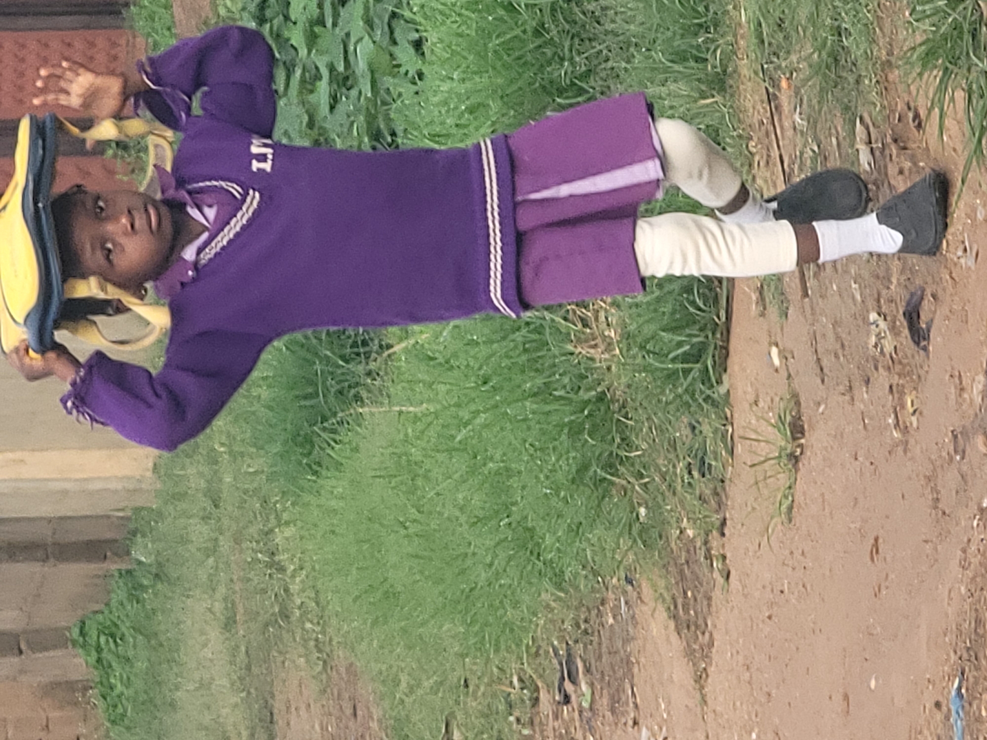 A young person, likely a child, is standing outdoors on a dirt path, wearing a purple school uniform. The uniform consists of a v-neck sweater with white trim and an emblem on the left chest that appears to read "LM". Below the sweater is a purple pleated skirt, worn over cream-colored leggings and white socks. The child is wearing dark shoes. The child is holding a yellow and blue backpack or bag on top of their head with both hands. They are looking towards the viewer with a neutral expression. The background features tall green grass and a concrete wall or building. The environment appears to be a street or pathway in a rural or semi-urban area. The location is identified as Barakin Akawo, Nigeria.