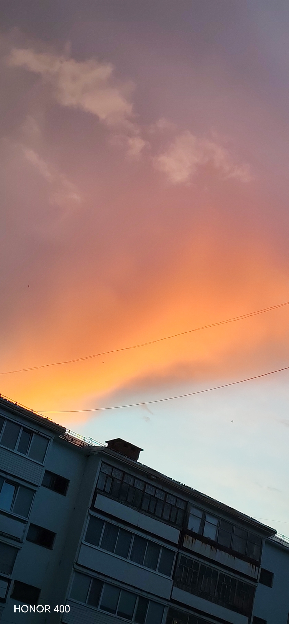 The image displays a vivid sunset or sunrise scene over a multi-story residential building in Noviy, Russia. The sky occupies the majority of the frame, showcasing a dramatic gradient of colors. At the very top, the sky is a muted purple-gray, transitioning downwards through soft pinks to a brilliant, fiery orange and yellow hue near the horizon line. Scattered clouds of various shapes and densities are illuminated by the low sun, enhancing the warm tones across the sky. Two thin, dark lines, resembling power or utility wires, stretch horizontally across the colorful sky, with a few tiny, dark specks, possibly birds, visible in the distance.
The lower third of the image features the upper sections of a light-colored building, likely an apartment block, viewed from a slightly upward and tilted angle. The building has numerous windows and balconies. Some balconies appear enclosed with glass, while others show darker, possibly open, interiors. Signs of wear and age are visible on the building's facade, including dark streaks or discoloration beneath some balconies. A dark, chimney-like structure is present on the roof. In the bottom-left corner of the image, the text "HONOR 400" is visible, indicating the camera or device used. There are no people visible, and no specific activity or event is taking place beyond the natural progression of day into night or night into day.