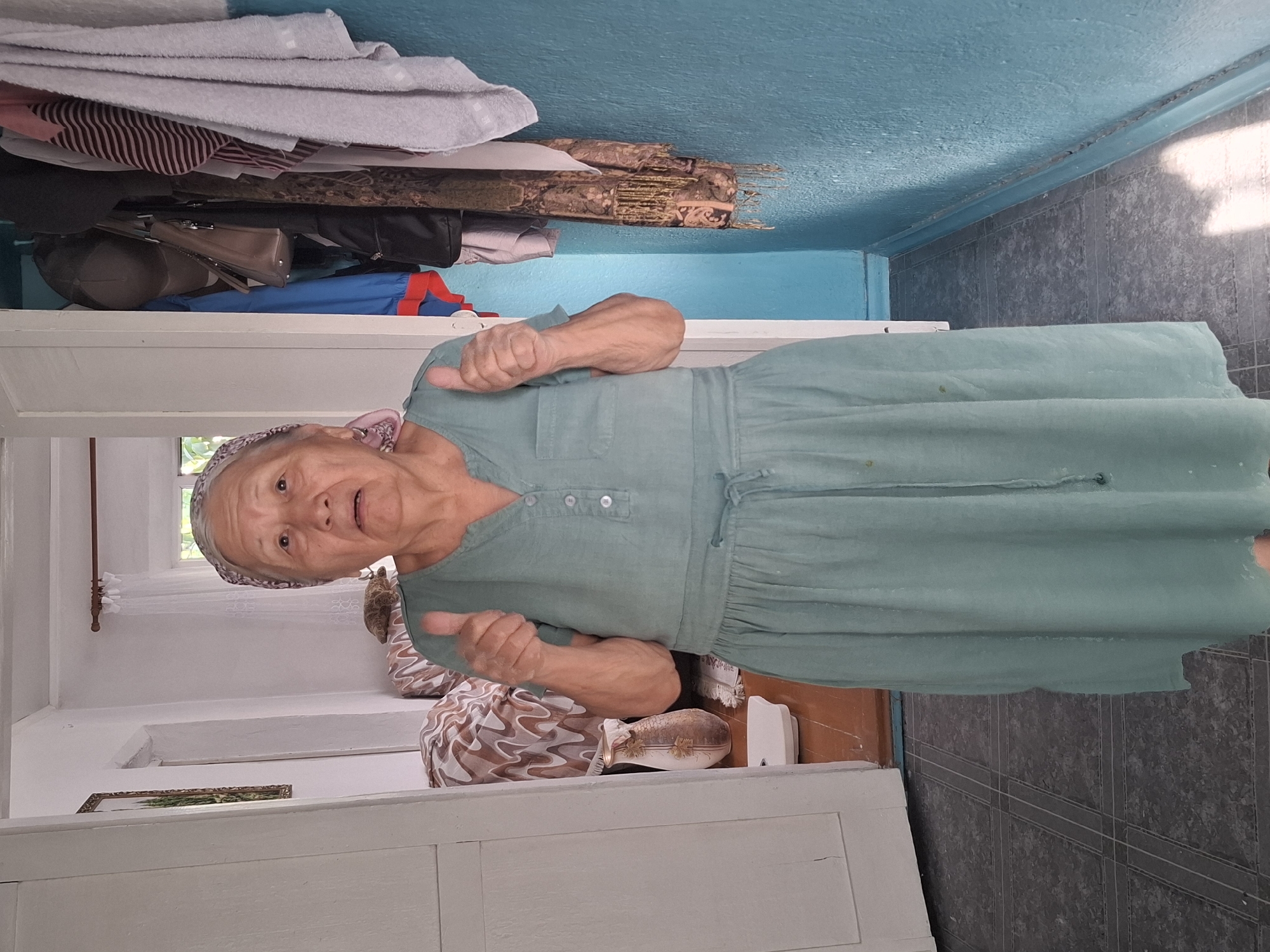 An elderly woman wearing a light teal dress and a headscarf stands in what appears to be a hallway, giving a double thumbs-up. The hallway has light blue walls and a dark tiled floor. To her left, a white door is ajar, revealing a glimpse of a room with a patterned sofa and a vase on a surface. To her right, a hook on the wall holds several items, including a dark bag, a striped towel, and a patterned scarf. The scene is lit by natural light, casting shadows on the blue wall. The image's composition suggests an indoor, possibly domestic, setting in Almaty, Kazakhstan.