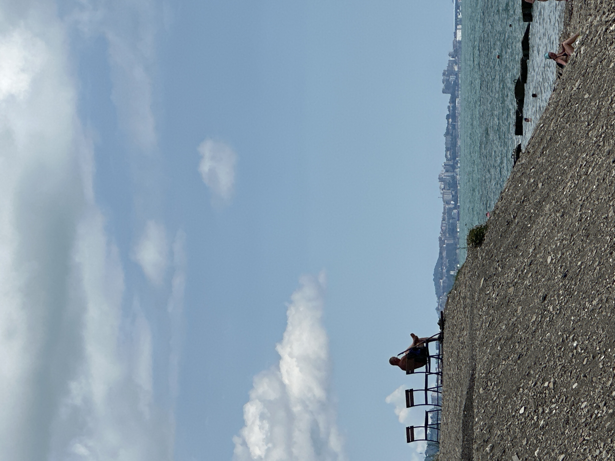 A person sits on a chair on a gravelly embankment overlooking the Black Sea. Two empty chairs are to the left of the person. In the distance, a city skyline is visible, likely Sochi, Russia, with buildings and a bridge. The sea is a turquoise color, and there are a few people visible in the water and on the shore in the distance. The sky is blue with scattered white clouds. The overall scene depicts a relaxed day at the beach with a view of the city.