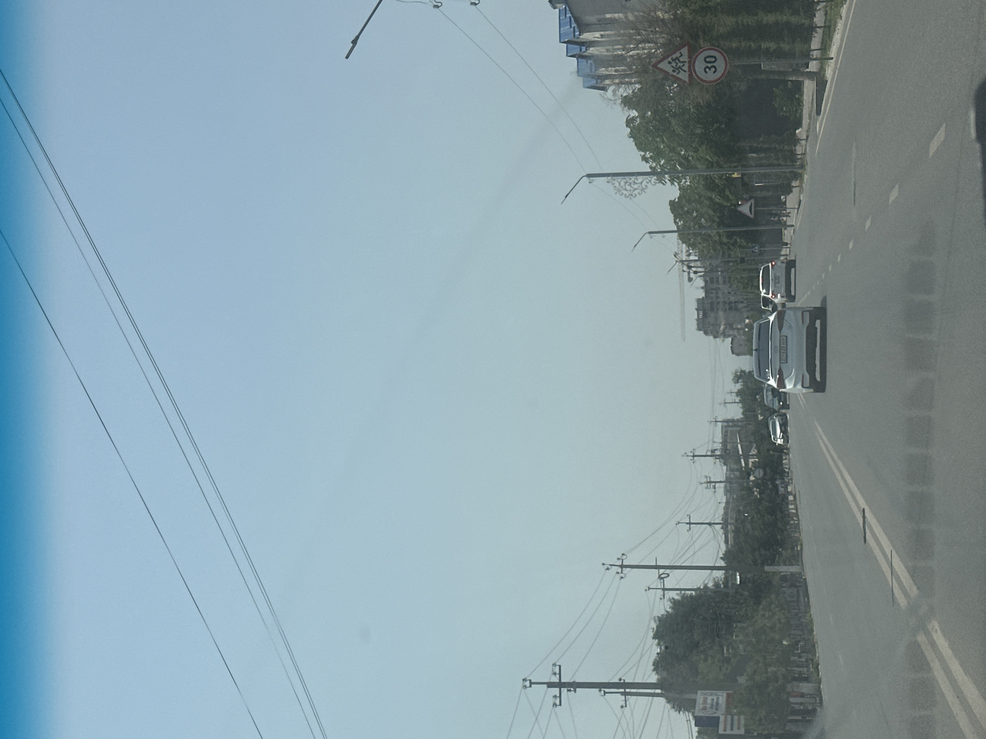 The image shows a street scene in Manas, Kyrgyzstan. The view is from a vehicle traveling down a paved road with double yellow lines in the center. On the right side of the road, there are tall trees, a sidewalk, and buildings with blue roofs. A traffic sign with the number "30" inside a red circle is visible, indicating a speed limit. Several cars are on the road, including a white car in the foreground with its license plate visible. On the left side of the road, there are power poles with numerous wires. In the distance, there are more buildings and construction cranes. The sky is clear and blue.
