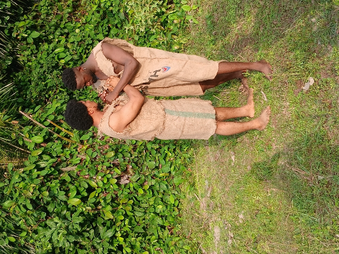 Two barefooted men, dressed in burlap sacks, lie on a grassy and leafy surface. They appear to be engaging in an altercation. The man on top has dark hair and a beard. The other man also has dark hair. Their bodies are positioned so they face each other amidst the verdant foliage.