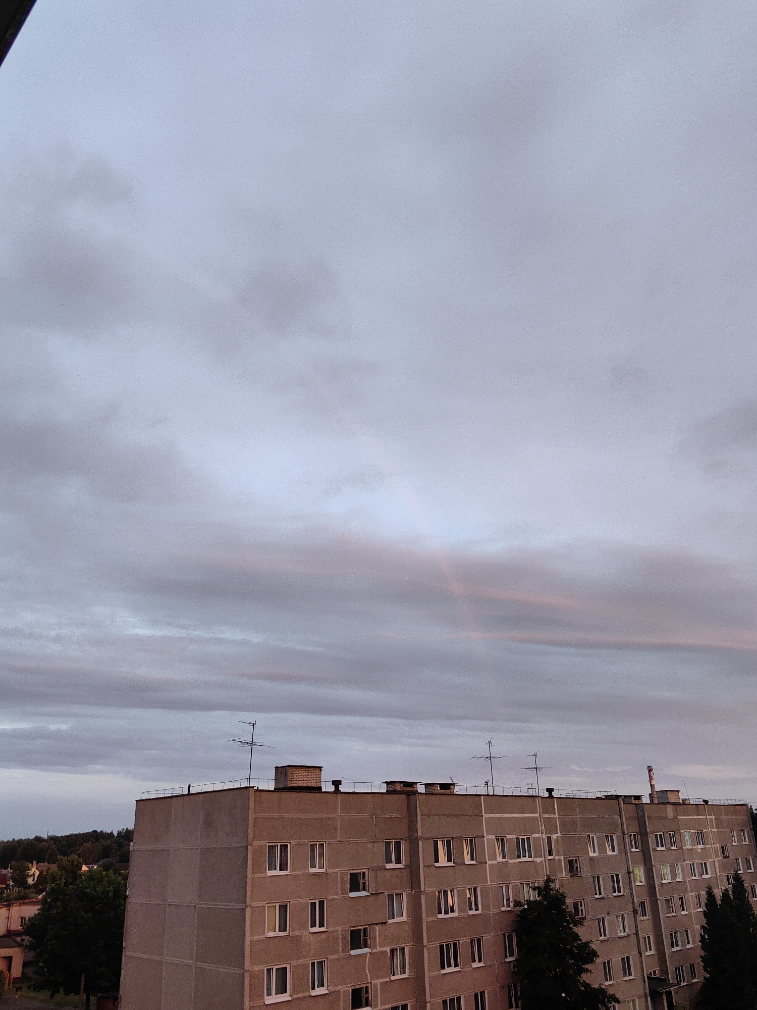 The image captures a section of a multi-story residential building in the city of Fanipal, Belarus, against a cloudy sky during what appears to be twilight. The building is constructed from concrete panels and features rows of windows, many of which have white frames and appear to be closed. The roof of the building is flat and has various antennae and chimneys. In the background, a small section of greenery, including trees, is visible to the left, with hints of other buildings in the distance. A faint rainbow is visible in the sky above the building, formed by the interplay of light and clouds. The overall scene is a typical urban residential landscape.