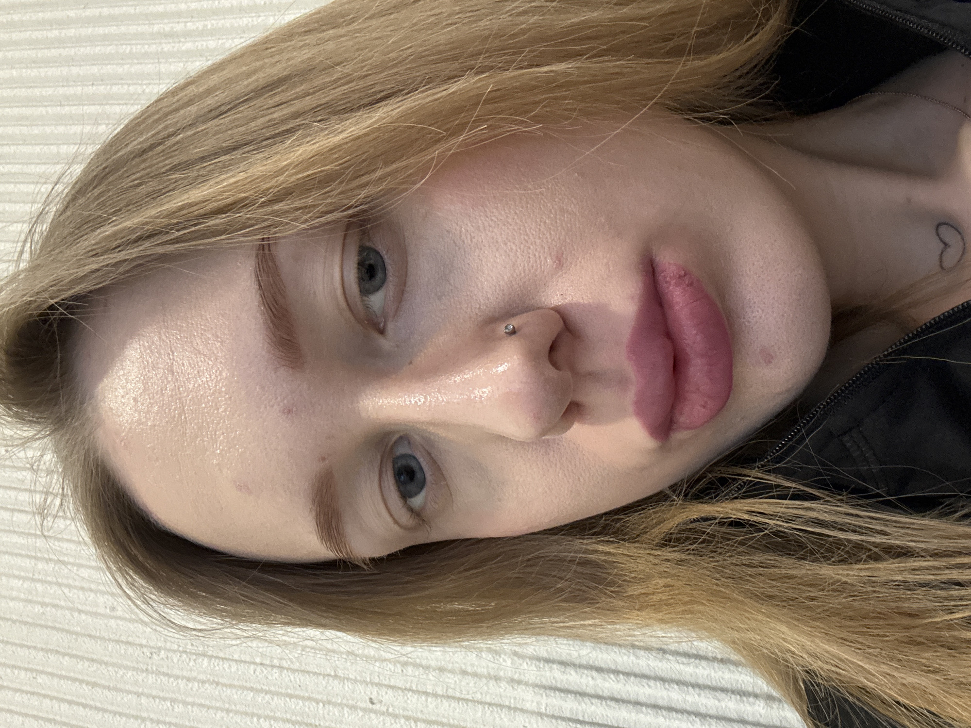 A close-up portrait of a fair-skinned person with blond hair. The person has light eyebrows, blue eyes, a nose piercing on the left side, and pink lips. The person's neck and part of a black jacket are visible in the lower right corner of the image. A small heart tattoo is visible on the neck. The background is a white textured surface. There is no evidence to determine if this image was taken in Ovoshchnoy, Russia.