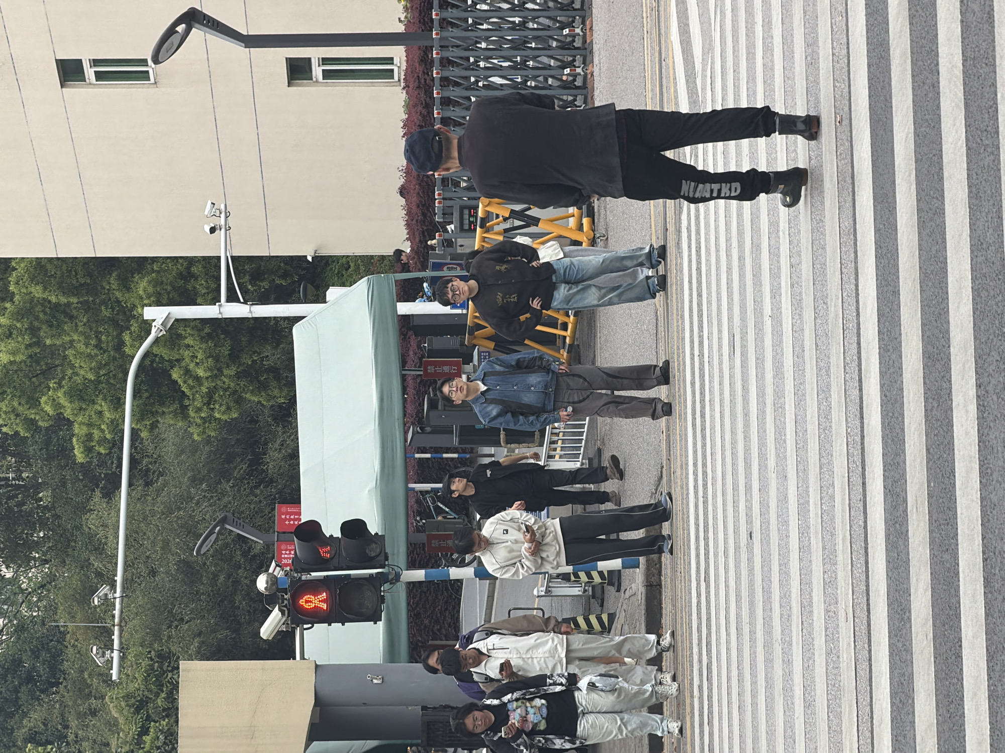 The image depicts a crosswalk scene in Jiangning District, China. The pedestrian crossing is marked with white stripes on a gray surface. A group of pedestrians are waiting at the crosswalk. The traffic light shows a red "X" symbol, indicating that pedestrians should not cross. A blue and white striped barrier is present near the traffic light. Various individuals are visible, including those wearing jackets and carrying bags. The area includes trees, buildings, and streetlights.