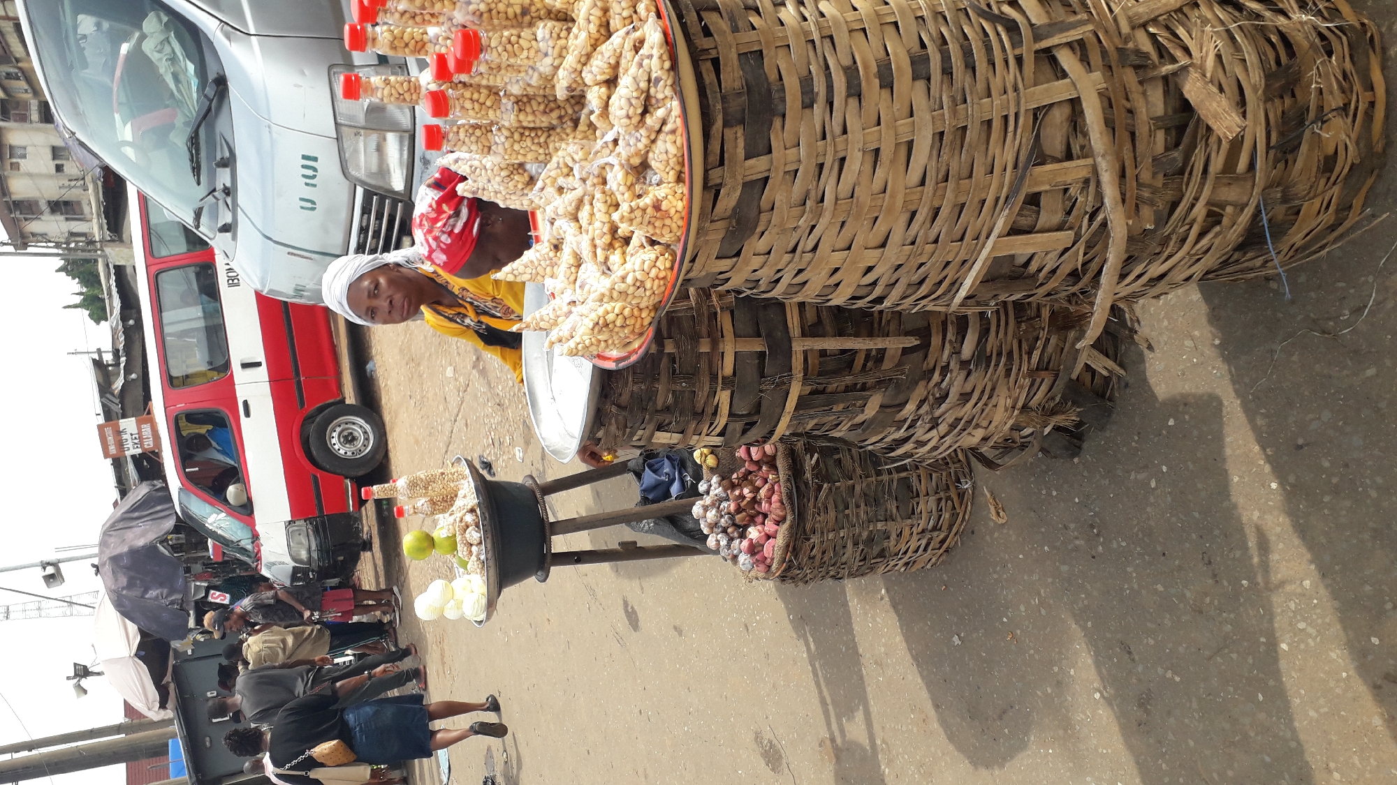 The image depicts a street market scene in Nkpor, Nigeria. In the foreground, two women are visible. One woman, wearing a yellow top and a white headscarf, is seated behind a display of goods. The other woman, whose head is covered with a red scarf, is partially visible behind her. The goods on display include baskets filled with nuts, some packaged in plastic bottles and others in clear plastic bags. To the left, there are also onions and what appear to be limes or lemons. Behind the women and their stall, two vehicles are parked on the street. One is a red and white van, and the other is a silver van. Several other people are walking in the background, and market stalls with awnings are visible. The ground is paved with concrete. The scene captures a moment of daily commerce in a bustling Nigerian city.