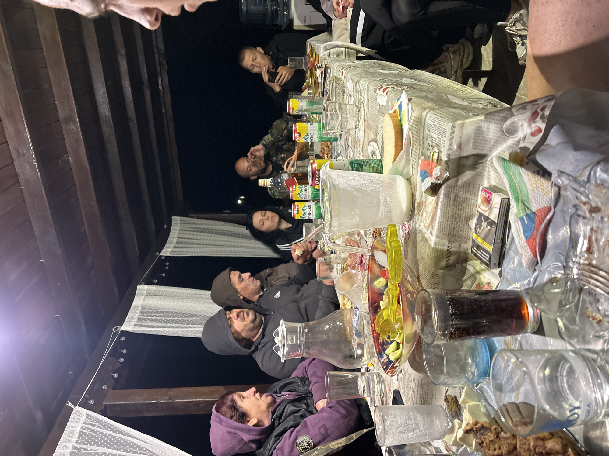 The image depicts an outdoor gathering around a table, possibly on a porch or patio, with a wooden overhead structure and draped white curtains. The setting appears to be evening or night, with artificial lighting present. Several people are seated around the table, engaged in conversation or eating.
On the table, there are various objects, including a glass pitcher, individual drinking glasses (some containing liquids), a bowl of salad, a plate with food items, cans of soda, a bottle of alcohol, and a pack of cigarettes. The table is covered with a patterned tablecloth.
In the foreground, a person's legs are visible, suggesting they are seated at the table and the photo is taken from their perspective. The people in the background are partially obscured by the table and other individuals.
The image does not contain any specific elements that would directly indicate a location in the city of Gabar, Bulgaria. The scene is generic and could represent a similar gathering in many locations.
