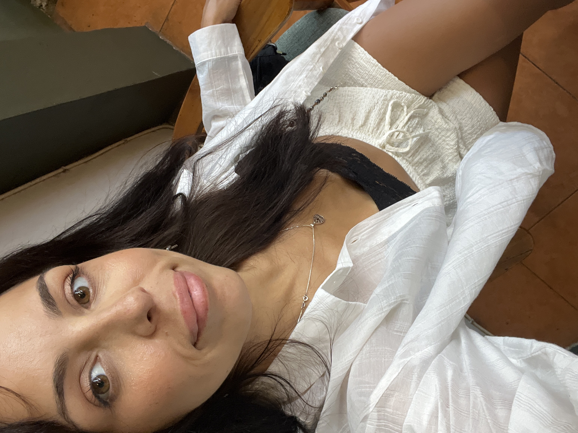 A woman with dark hair and tan skin is reclining indoors, wearing a white, long-sleeved, button-down shirt over a black top and white shorts. She has a silver necklace with a pendant. The floor is tiled in brown. The location is potentially Kuta Selatan, Indonesia.