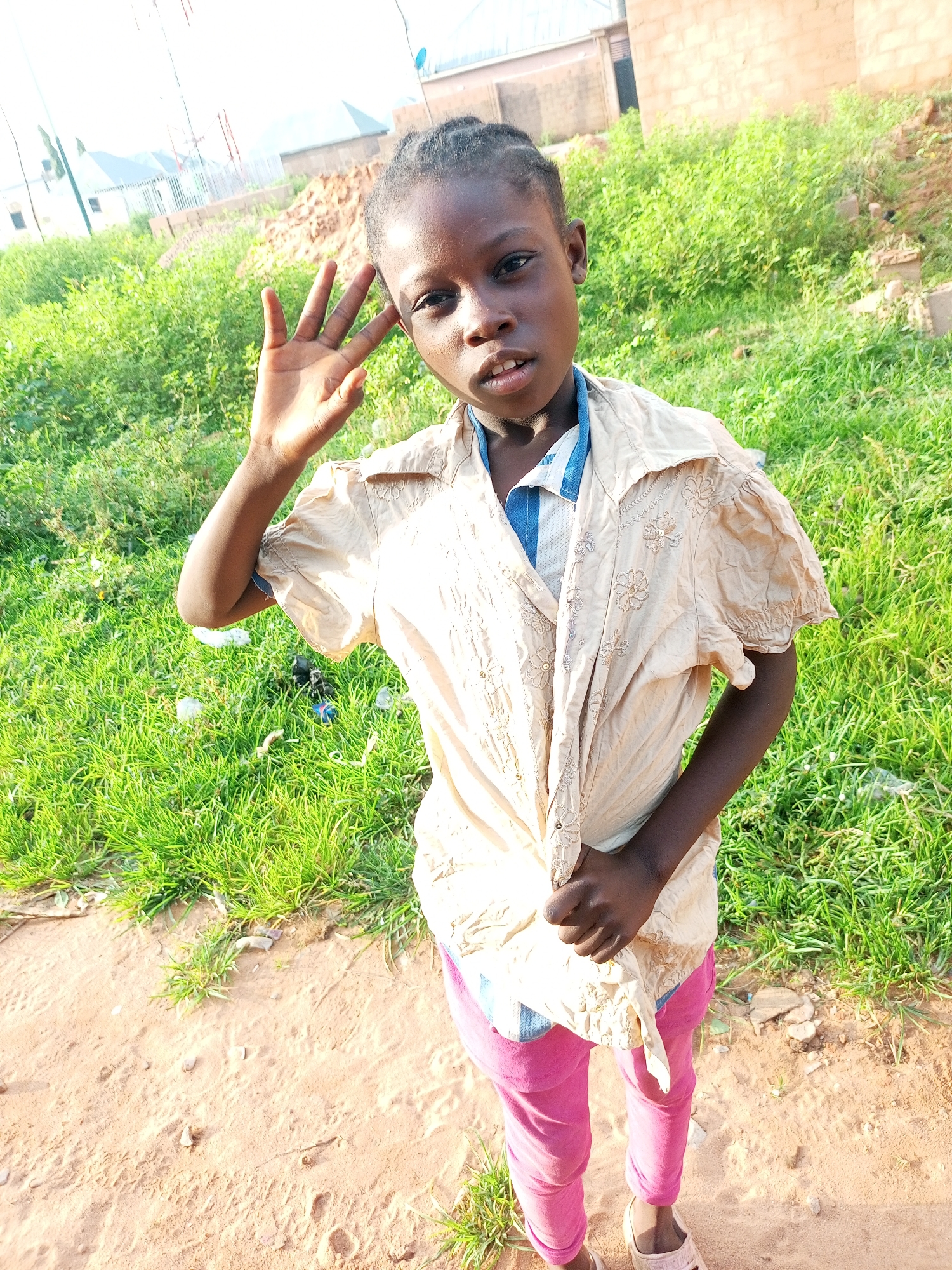 A young Black girl, appearing to be of school age, stands outdoors in what seems to be an undeveloped or construction area in Tempure, Nigeria. She is facing forward, looking directly at the camera. Her right hand is raised near her face, with her index and middle fingers extended in a peace sign. She has dark skin, braided black hair pulled back, and her mouth is slightly open as if speaking or posing.
She is wearing a light tan, collared shirt with short, puffed sleeves. The shirt has a pattern of embroidered flowers and is loosely tied at the waist. Underneath, she wears a blue and white striped collared shirt. Her pants are bright pink, and she has on light-colored sandals.
The environment behind her is filled with lush green grass, some scattered debris, and in the background, there are unfinished or partially constructed buildings. The ground in front of her is sandy and bare, with small stones and patches of grass. The lighting suggests it is daytime, possibly late afternoon, with the sun casting distinct shadows. The overall impression is of a candid portrait taken in a rural or developing urban setting.