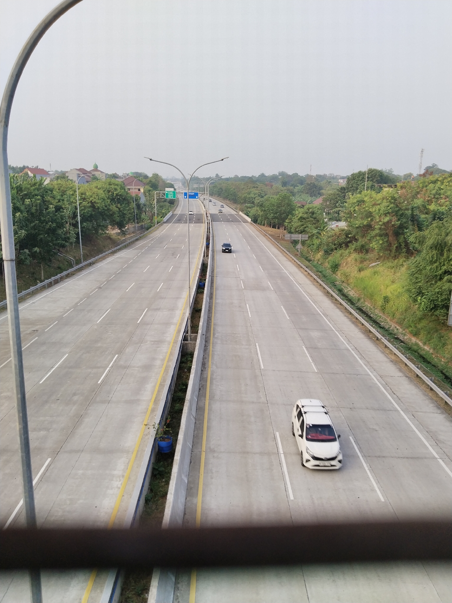 The image shows an elevated view of a multi-lane highway in Depok, Indonesia, likely an expressway. The highway is paved with concrete and marked with white lane dividers and yellow edge lines. Two lanes are visible in each direction of travel. The sky is overcast, suggesting it is either early morning or a cloudy day. Lush green trees and vegetation line the sides of the highway, with some residential buildings and structures visible in the distance.
Several vehicles are present on the highway. In the foreground, on the right-hand side of the highway, a white multi-purpose vehicle (MPV) is traveling in the rightmost lane. Further down the highway, on the left-hand side, a dark-colored car is also traveling. Other vehicles can be seen as small dots in the distance, indicating traffic flow.
Streetlights are mounted on tall poles along the sides of the highway, and signage is visible, including green directional signs. In the median area between the two directions of the highway, there is a drainage channel with some vegetation and a blue barrel with a small plant. The overall impression is of a well-maintained and functional transportation infrastructure within a semi-urban or suburban environment.