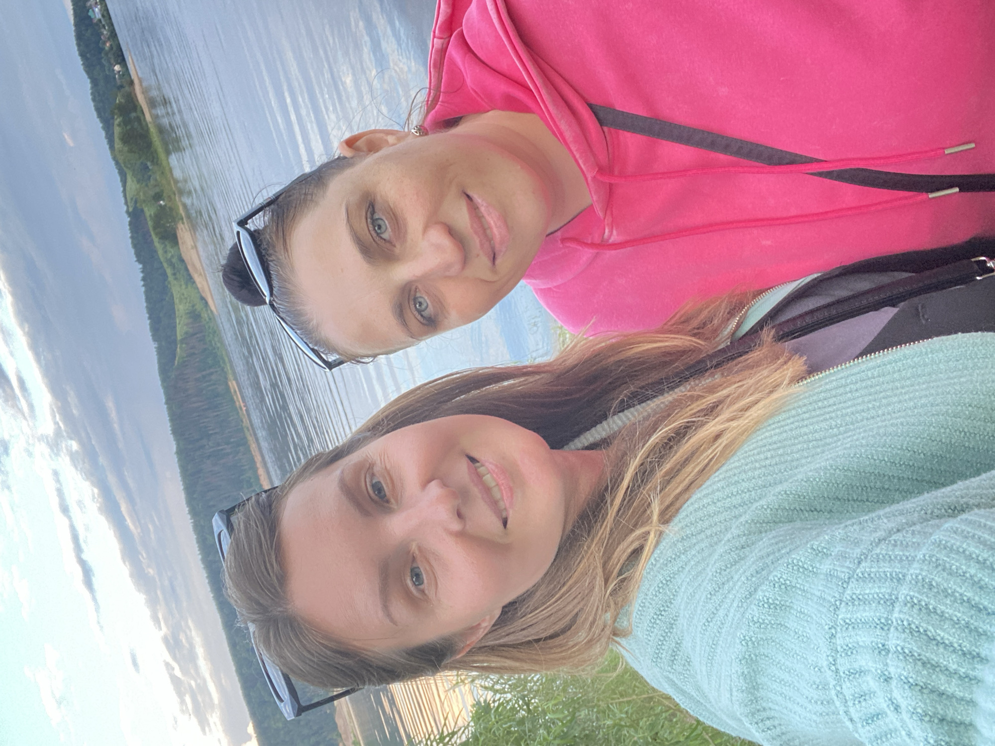 This is a selfie of two women. The woman in the foreground has blonde hair and is wearing a light blue knitted sweater. She is smiling. The woman in the background has dark hair pulled back and is wearing a bright pink hooded sweatshirt. She is also smiling and looking at the camera. Both women are wearing sunglasses pushed up on their heads.
They are outdoors, next to a body of water that appears to be a river or lake. The background shows a green, tree-covered bank and a cloudy sky. The lighting suggests it could be late afternoon or early evening. The overall mood is relaxed and happy. The location is identified as Noviy, Russia. There is no visible text in the image.