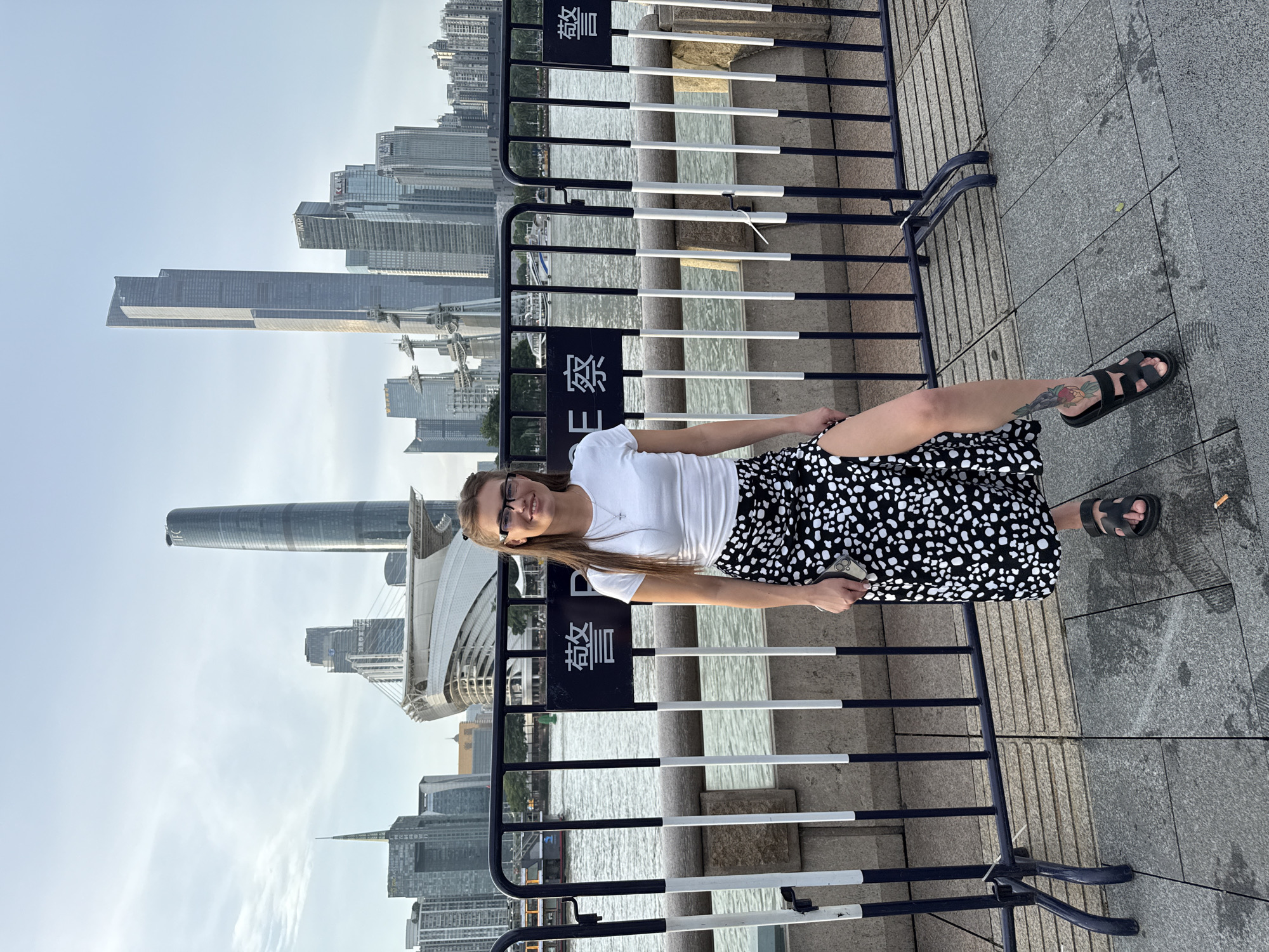 A young woman with brown hair and glasses stands in front of a fence with Chinese characters that translate to "Police." She is wearing a white t-shirt and a black and white spotted skirt with a high slit. She is also wearing black sandals. In the background, there is a river and a cityscape with tall buildings. The image appears to be taken in Haizhu District, China, likely along the Pearl River. The woman is smiling and looking at the camera.