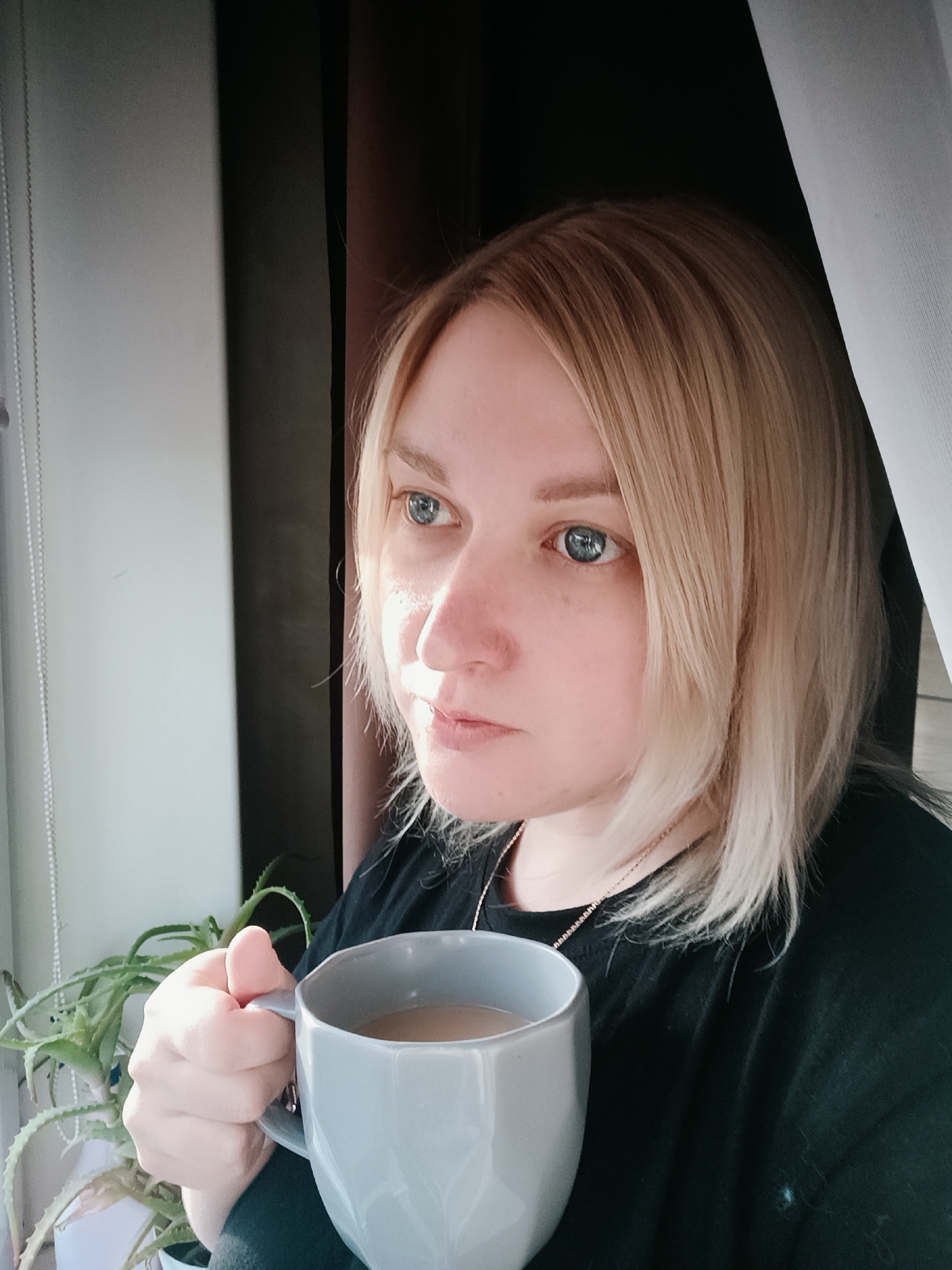 A light-skinned woman with shoulder-length blonde hair and striking blue eyes is the central subject of this indoor portrait, taken in Chaikovsky, Russia. She is wearing a dark, possibly black, long-sleeved top and a delicate necklace. She holds a grey, faceted mug containing a light brown beverage, likely coffee or tea with milk, in her right hand. Her gaze is directed pensively out a window to her left, suggesting a moment of quiet reflection.
The scene is an indoor setting, with the woman positioned close to a window. White vertical blinds are partially visible to her left, and bright natural light streams in, illuminating the left side of her face. A green succulent plant, resembling aloe vera, sits in a white pot on the windowsill. Dark curtains are visible in the background to her right. The strong natural light indicates it is daytime. Her expression is calm and contemplative. There is no visible text in the image.