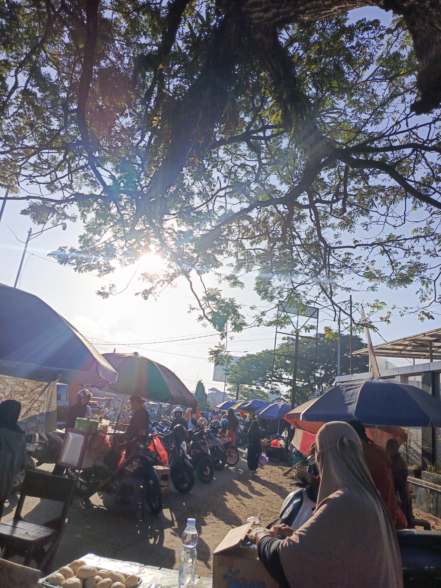 A low-angle shot captures an outdoor market scene under a sprawling tree canopy in Kendari, Indonesia. The sunbeams pierce through the leaves, creating lens flares. In the foreground, a tray of small, round, baked goods sits on a table. To the right, a person wearing a light brown hijab is seated on a motorcycle, holding a cardboard box. Behind them, other market stalls with colorful umbrellas and more motorcycles are visible. A woman in a black hijab is walking in the background. The ground appears to be dirt and sand. The overall atmosphere suggests a bustling morning market.