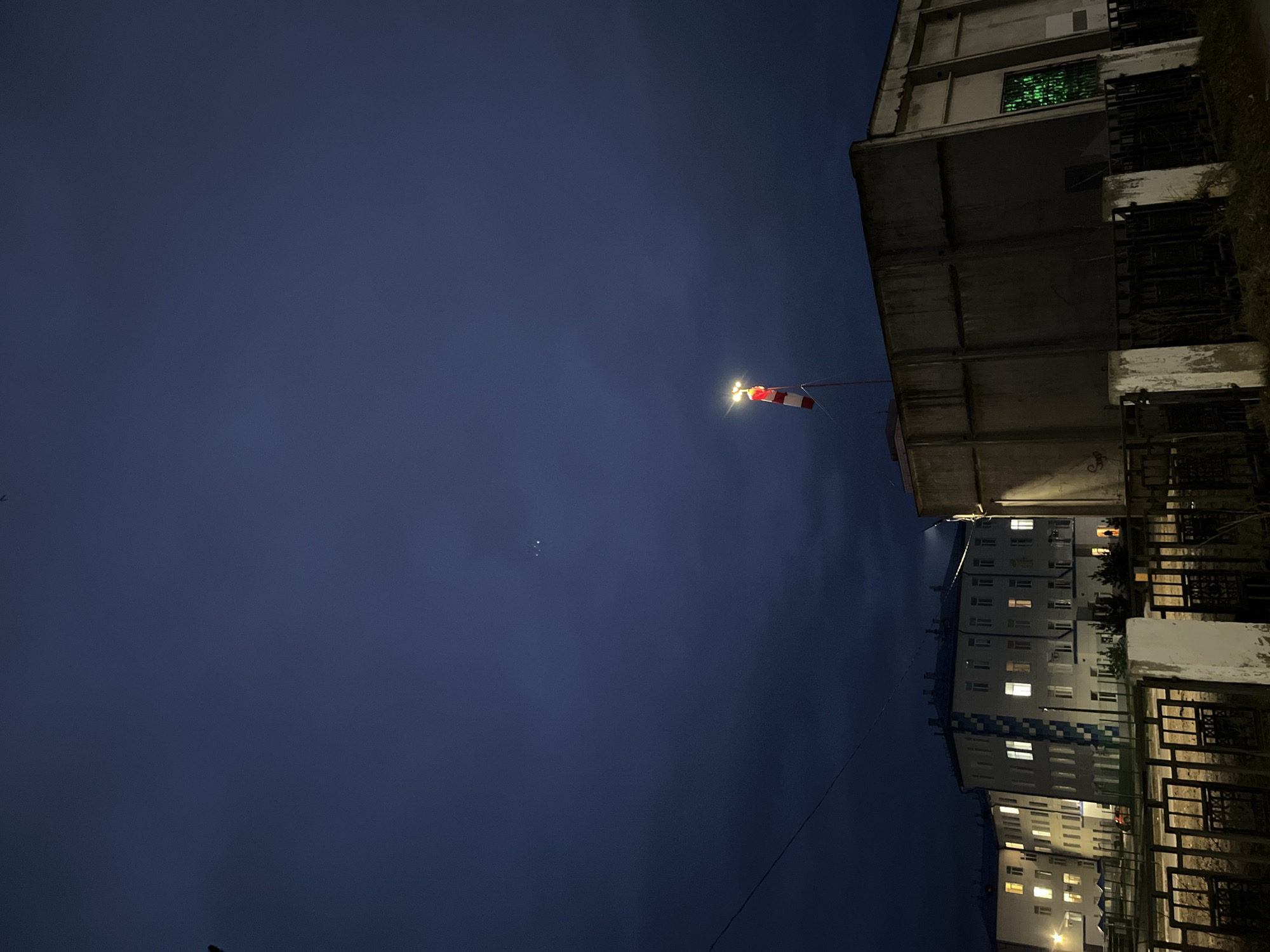 The image captures a nighttime urban scene in Chaikovsky, Russia, characterized by a deep blue sky and illuminated structures. In the upper right quadrant, a red and white striped windsock is prominently displayed on a pole, brightly illuminated by an external light source, indicating wind direction. To its left, a small, bright white point of light is visible in the dark sky.
On the right side of the frame, a stark, multi-story concrete building with a weathered appearance dominates the upper section. One of its windows on the right facade emits a striking, bright green light. Dark metal railings or grates are visible on the lower levels of this building.
Below this and extending across the bottom right, a large residential apartment building is seen. Many of its windows are lit, suggesting habitation. The building features a light-colored facade with a distinctive diagonal blue pattern on one section. A dark, decorative metal fence occupies the immediate foreground on the right, separating the viewer from the residential block. A thin dark wire crosses diagonally from the lower right towards the center-left, passing above the apartment building. The scene conveys a quiet, late-night atmosphere with artificial light sources providing the main points of focus. No text is visible in the image.