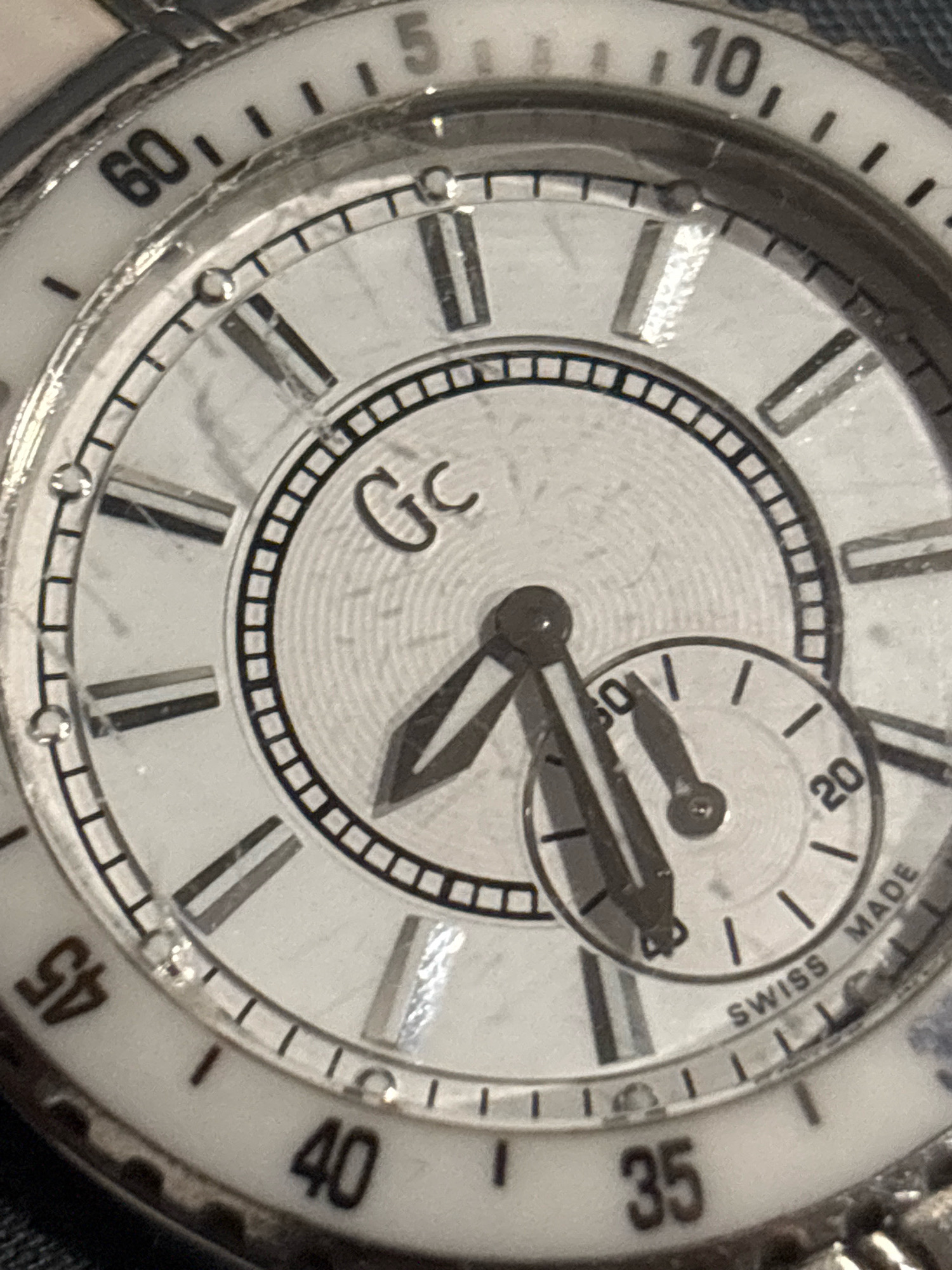 The image shows a close-up of a watch face. The watch has a white dial with Roman numerals for hours 1 through 12 on the outer rim, marked in increments of 5. The numbers 5, 10, 35, 40, and 45 are clearly visible. There are also smaller minute markers between the hour markers. In the center of the dial, the logo "GC" is visible. There are two hands on the watch, a longer hour hand and a shorter minute hand, both pointing towards the 8 o'clock position. A smaller sub-dial is located in the lower right quadrant of the main dial, with a smaller hand pointing slightly past the 25-minute mark, and the numbers 20 and 30 visible. The text "SWISS MADE" is also present on the lower right edge of the main dial. The watch appears to be a wristwatch with a metal casing. The background is dark and out of focus. The image does not contain any information that would indicate its location in Ihtiman, Bulgaria, or any other city.