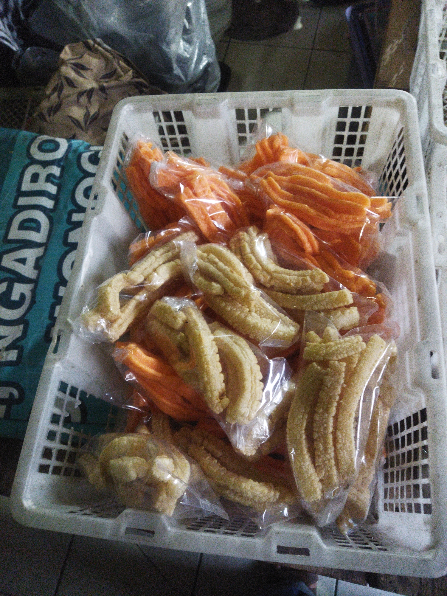 The image shows a close-up, overhead view of a white plastic basket filled with packaged snacks. The basket is positioned on a dark, textured surface, likely a floor or table. Inside the basket, there are two types of snacks. One type is a light yellow, ridged snack, packaged in clear plastic. The other type is an orange, elongated snack, also in clear plastic. The snacks are piled high, filling the basket. To the left of the basket is a portion of a blue and white patterned bag or cloth, with text that appears to be in a foreign language. In the background, out of focus, are other items including plastic bags and what appears to be a patterned fabric. The scene suggests a market or store setting, possibly in Wonogiri, Indonesia, given the context of the request, though the image itself does not provide definitive location details. There are no people visible in the image.