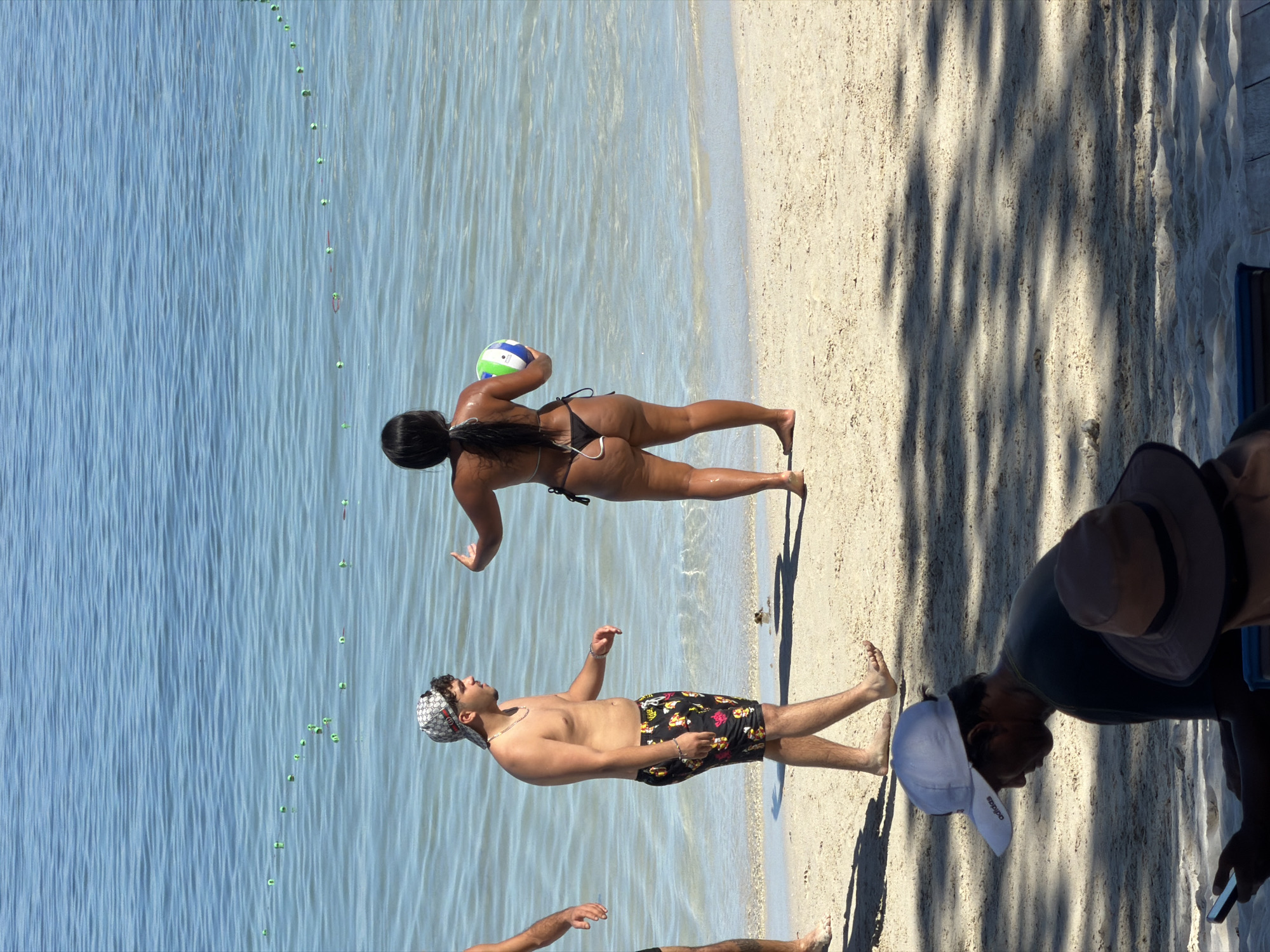 The image depicts a sunny day at a beach. In the foreground, two individuals are partially visible, seated in the shade of trees, one wearing a white cap and looking at a phone, the other wearing a wide-brimmed hat. In the mid-ground, a man in floral swim trunks and a patterned baseball cap walks along the sand. Behind him, a woman in a black bikini stands by the water, holding a striped ball. The ocean is calm and clear, with a line of buoys visible in the water. The beach appears to be made of light-colored sand. The scene is consistent with a beach environment in Baan Chaweng, Thailand, known for its popular beaches.