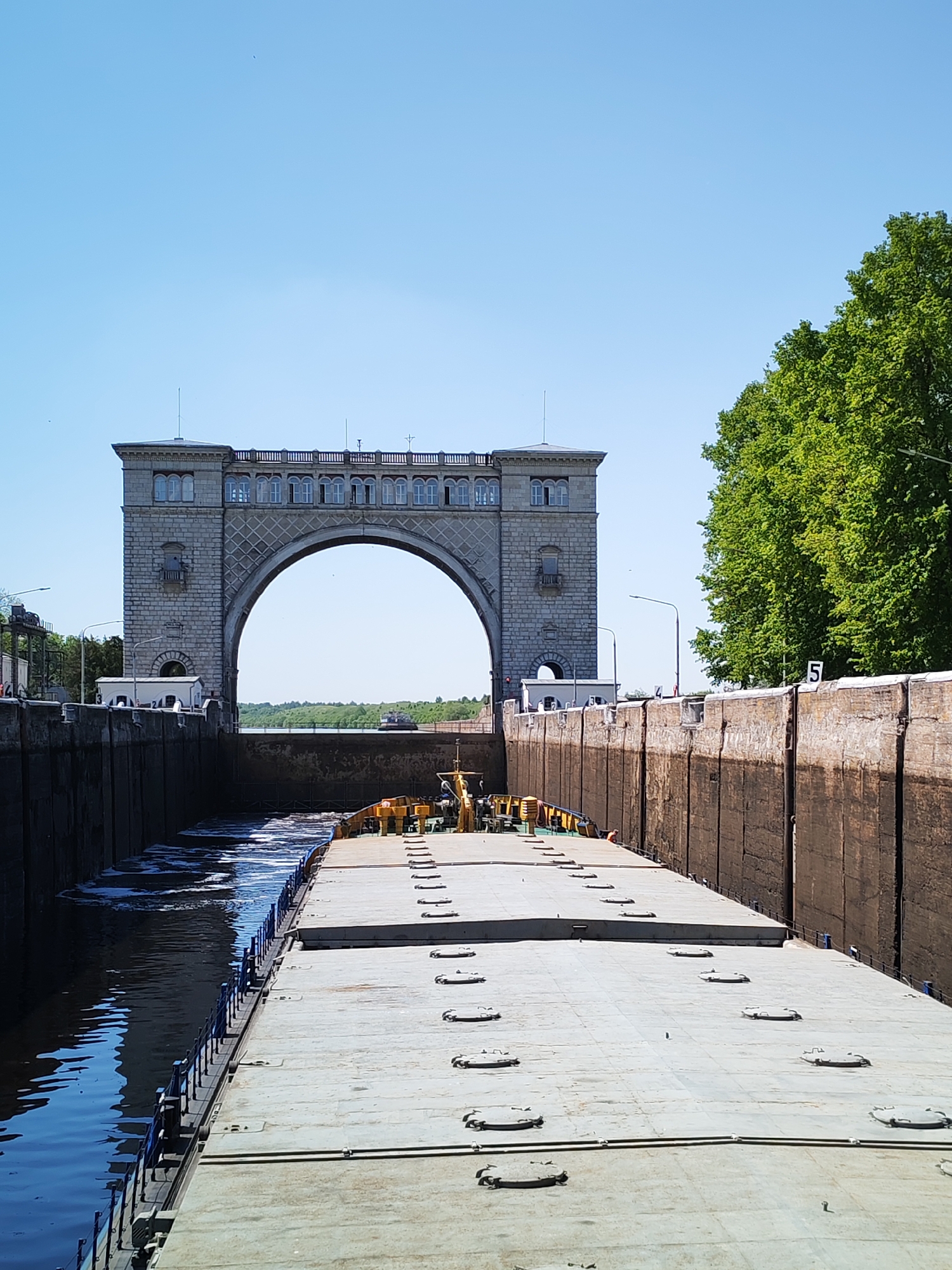 A barge is positioned in a lock on the Volga-Don Canal, Russia. The lock walls are constructed from concrete and numbered "5" on the right side. The arch of the lock is visible in the background. Trees line the lock on the right, and open sky fills the upper portion of the image.