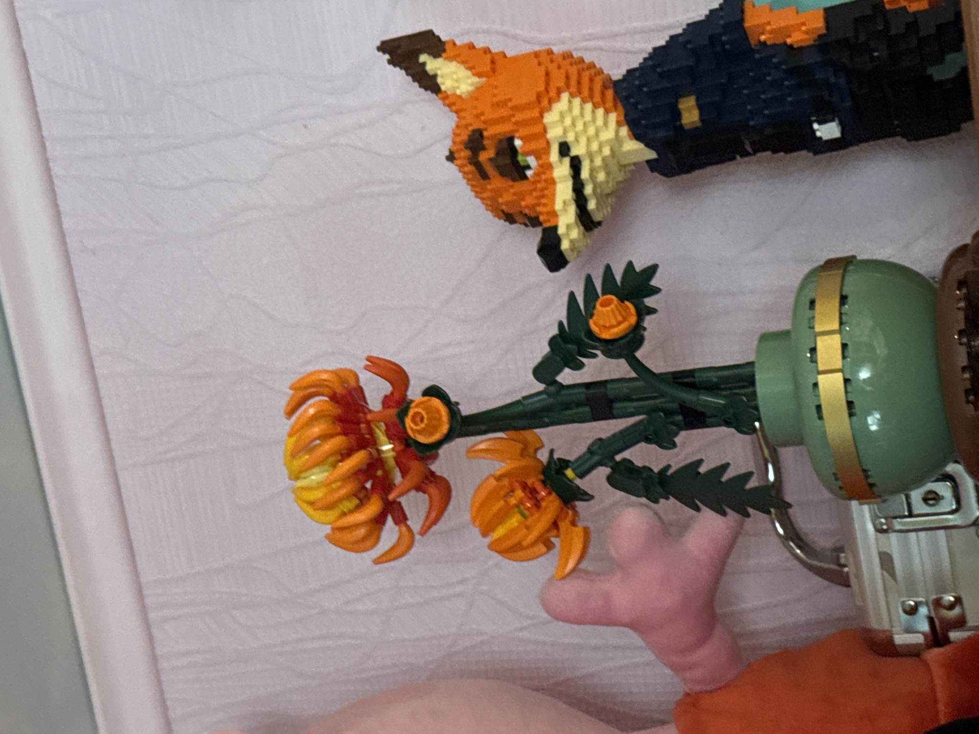 In an indoor setting, a LEGO model of a fox character with orange fur and a dark blue uniform stands to the right of the frame. In the center, a LEGO floral arrangement features orange and yellow flowers with green stems and leaves, placed in a green vase with a gold band. To the left, a pink plush toy hand is visible, along with a portion of an orange plush toy. A silver suitcase with clasps is positioned in the foreground, below the floral arrangement and to the left of the fox. The background consists of a textured, light-colored wall.