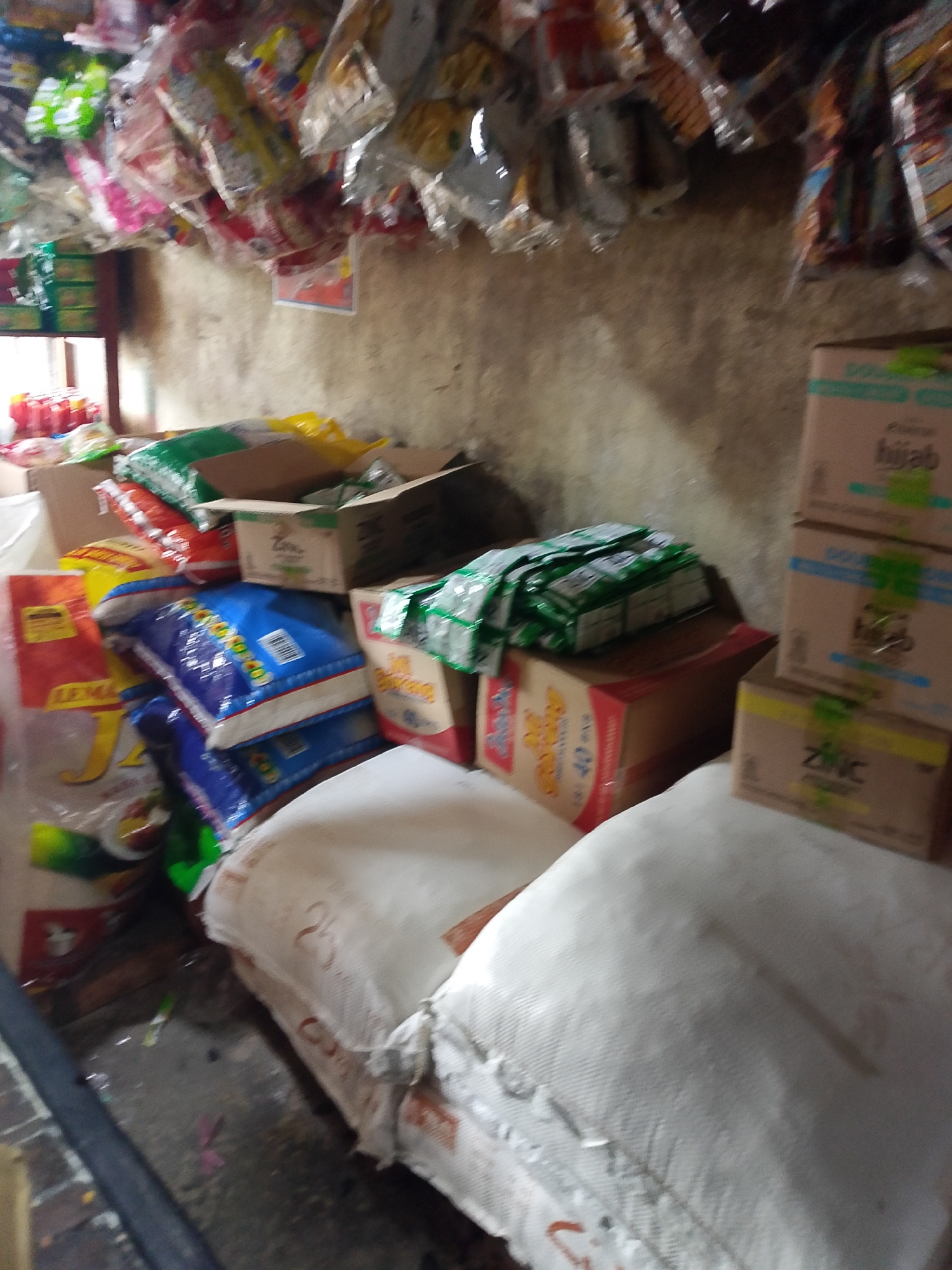 The image depicts a dimly lit, cluttered interior space, likely a small shop or storage area, in Palembang, Indonesia. The scene is dominated by stacks of boxed goods, plastic-wrapped products hanging from above, and large sacks or bags on the floor. Prominently displayed are various packaged food items, including what appears to be rice in blue and yellow packaging, as well as small green packets stacked on a red and yellow box. Several cardboard boxes are visible, some with labels in Indonesian such as "hijab" and "ZinC". The walls are rough and textured, suggesting a simple building structure. The overall impression is one of a commercial space filled with inventory.