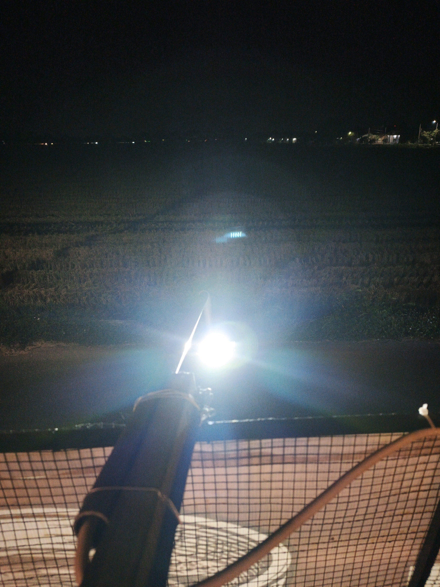 The image depicts a night scene in Ketegan, Indonesia. In the foreground, a flashlight is aimed forward, illuminating a pathway and a field. The flashlight is attached to a cylindrical object with a dark casing, secured by a strap. A wire mesh fence runs horizontally across the bottom of the image. In the background, a dark field stretches towards a distant horizon dotted with faint lights, suggesting the presence of buildings or a settlement. The overall atmosphere is one of darkness, punctuated by the bright beam of the flashlight.