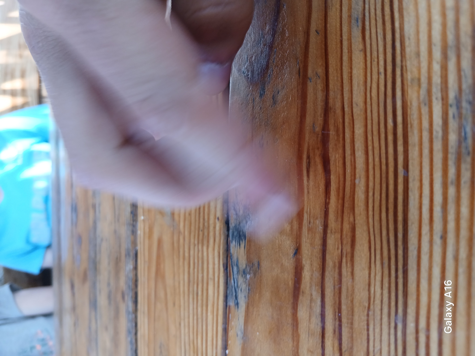 The image shows a close-up of a hand partially obscuring a wooden surface. The wooden surface has distinct vertical grain patterns and appears to be textured. The hand is blurred, suggesting movement or a shallow depth of field. To the left of the hand, a person wearing a bright blue shirt is visible, also out of focus. The background suggests an outdoor setting with some bright light, possibly indicating daytime. The overall scene is casual and appears to be taken in a natural environment. Visible text in the lower right corner reads "Galaxy A16". The location context is Ustyvytsia, Ukraine.