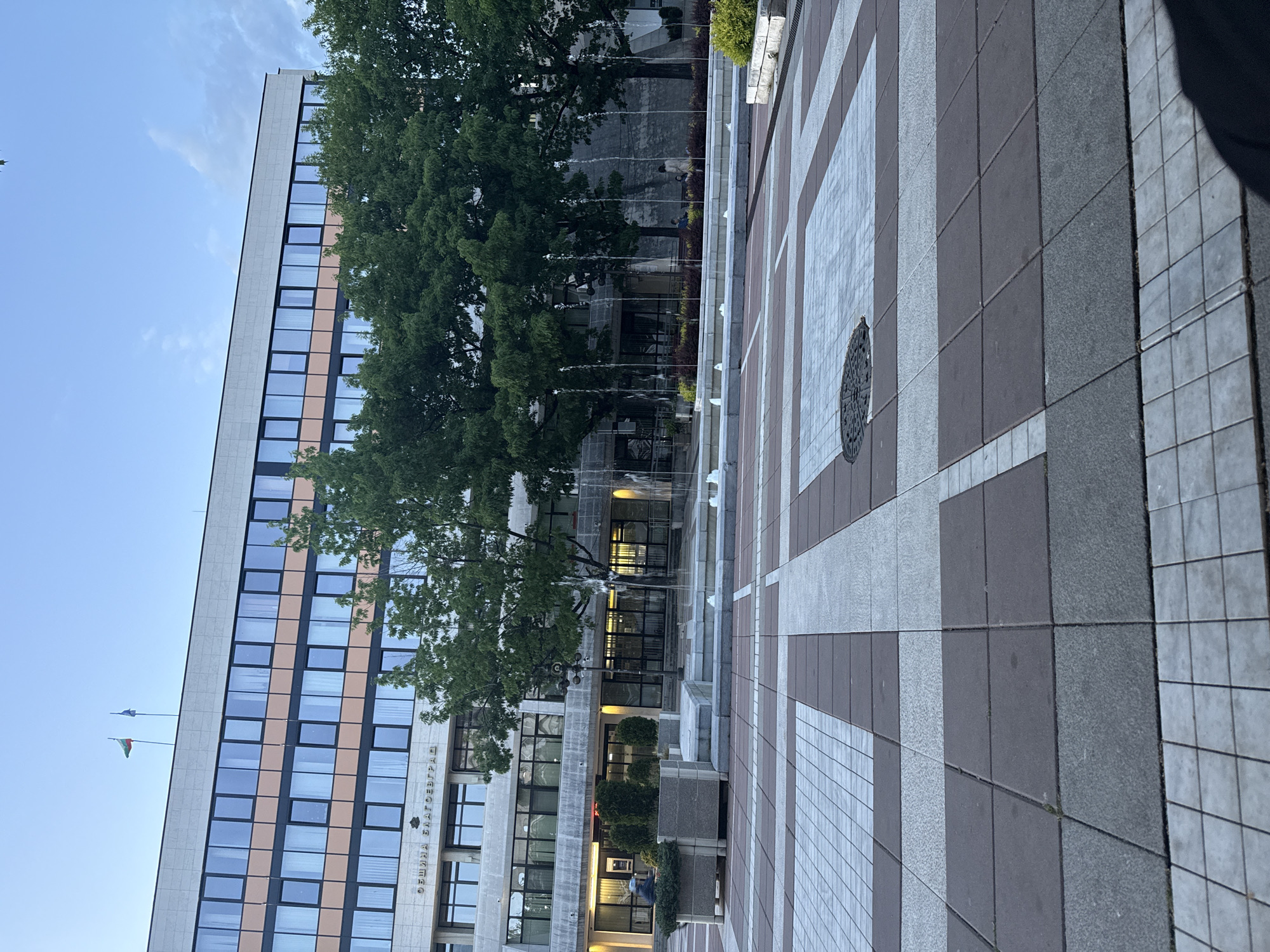 The image shows the Municipality of Blagoevgrad in Bulgaria. The building is tall, with numerous windows. Flags fly atop the building. In front of the building, there is a fountain and a paved area with a pattern of light and dark gray squares. Trees are interspersed in the scene. The sky is visible in the background.