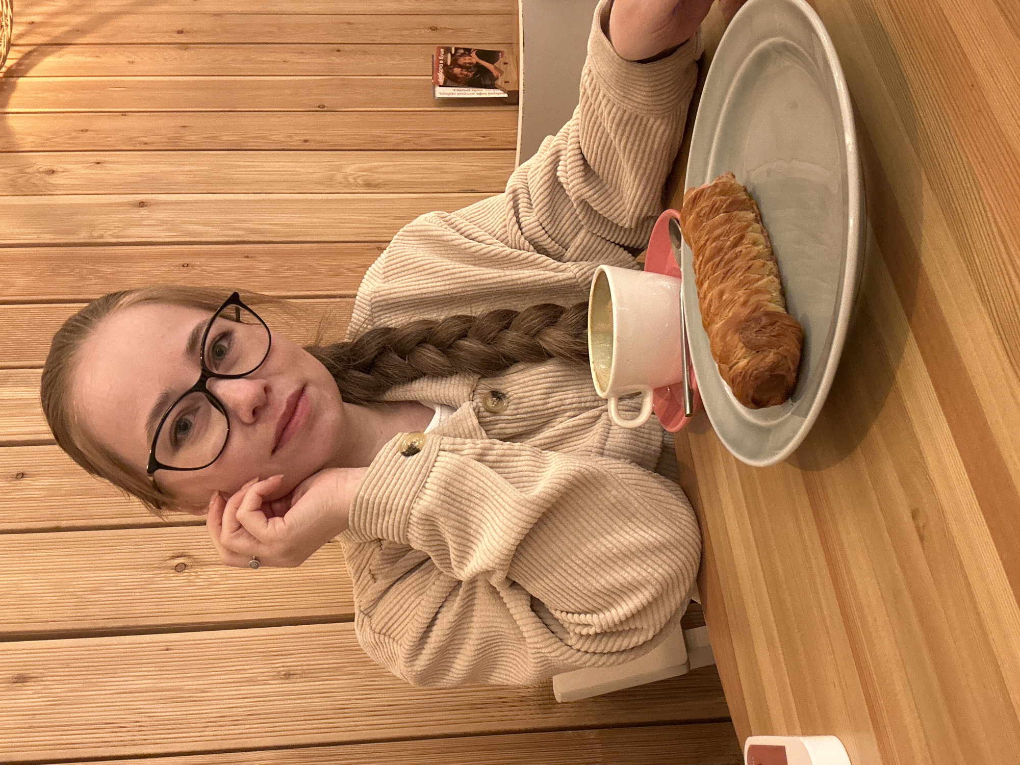 This image, captured in Saint Petersburg, Russia, features a young woman seated at a wooden table with matching wooden paneled walls as a backdrop. She has light brown hair styled in a long braid that drapes over her shoulder and across her chest. She wears black-rimmed glasses and a light beige, corduroy jacket or shirt. Her left hand, adorned with a ring, supports her chin as she looks towards the camera with a calm, relaxed expression.
On the table in front of her, there's a light gray oval plate holding a golden-brown braided pastry. Beside the plate, a white mug rests on a pink saucer, suggesting she is enjoying a beverage. The table and wall surfaces consist of light-colored wooden planks, creating a warm, cozy atmosphere, likely in a cafe or a similar casual indoor setting.
In the background, a small framed picture or card is affixed to the wooden wall. Clearly visible text on it, written in Cyrillic, reads "Свобода и душе" (Freedom and soul). Other, less legible text is present below it. The woman appears to be relaxing, possibly taking a break to enjoy her snack and drink.