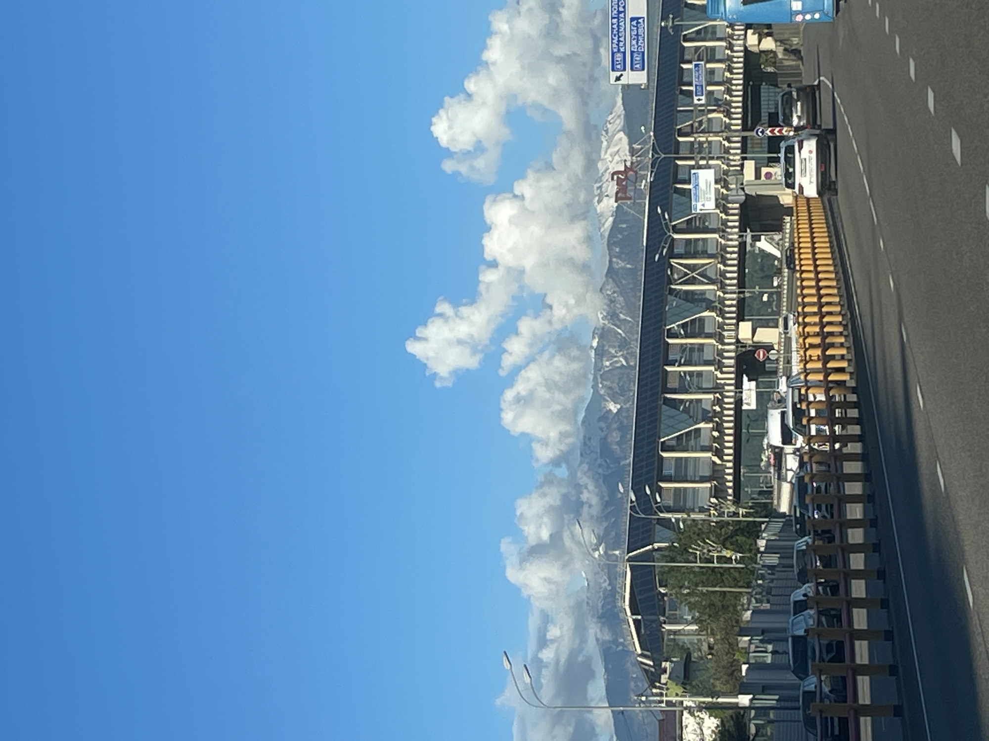 A landscape-oriented photograph displays a highway in Sochi, Russia, flanked by a mountainous skyline on the horizon. The sky occupies a significant portion of the frame, exhibiting a gradient from deep to pale blue, interspersed with cumulus clouds. Parallel to the highway, there is a series of modern architectural structures, possibly commercial or residential buildings. The road itself is composed of asphalt with double white lines, indicative of lane markings. Along the road's edge, a yellow and grey barrier separates the highway from the adjacent buildings. Cars are seen parked along the highway. Street lights and traffic signs are visible along the highway.