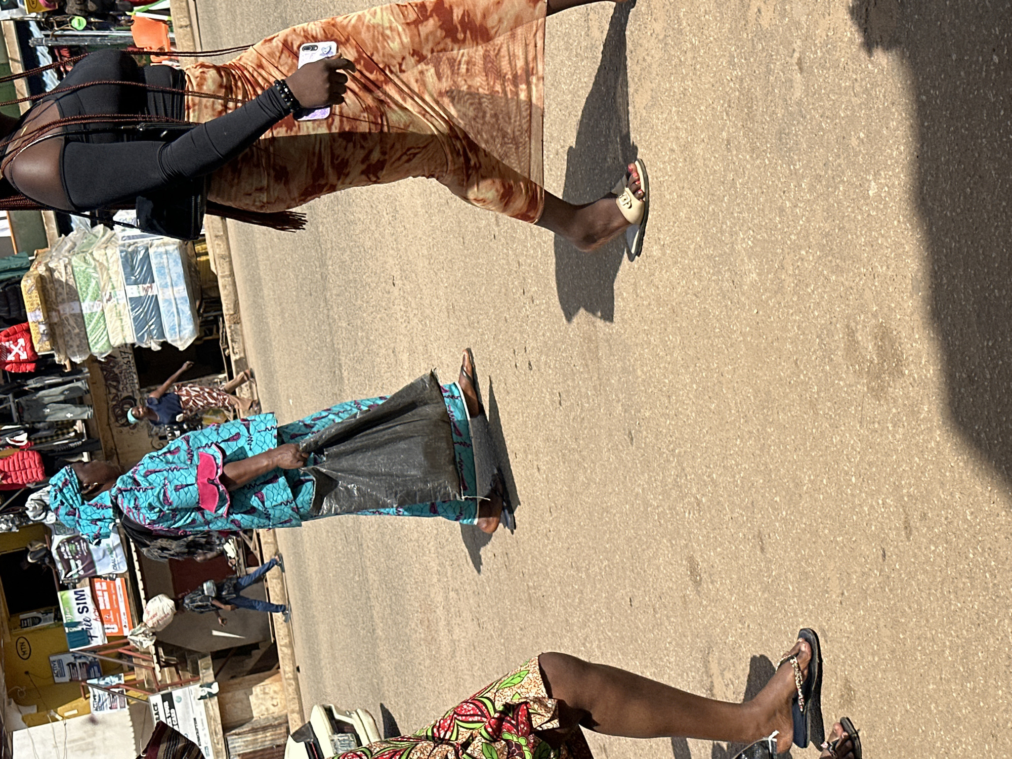 The image depicts a street scene in Bukuru, Nigeria, under daylight conditions. In the foreground, the legs and feet of three individuals are visible as they walk on an asphalt road. The person on the far left, partially obscured, wears a colorful patterned garment and black flip-flops with decorative gold accents. To their right, a person is seen from the knees down, dressed in a vibrant blue and teal traditional African outfit with pink trim, carrying a large black plastic bag in their right hand and wearing dark sandals. The third person, on the far right, is seen from the mid-thigh down, wearing a long, flowing orange and brown tie-dye skirt and light-colored slides.
In the background, stalls line the street, displaying various goods, including stacked mattresses, clothing on racks, and signage. One sign prominently features the "MTN" logo. Several people are visible in the middle ground, including a person wearing a blue shirt and jeans walking in the distance, and another person in the background wearing a patterned skirt and blue top. A white vehicle is parked on the left side of the street. The environment appears to be a bustling urban marketplace.