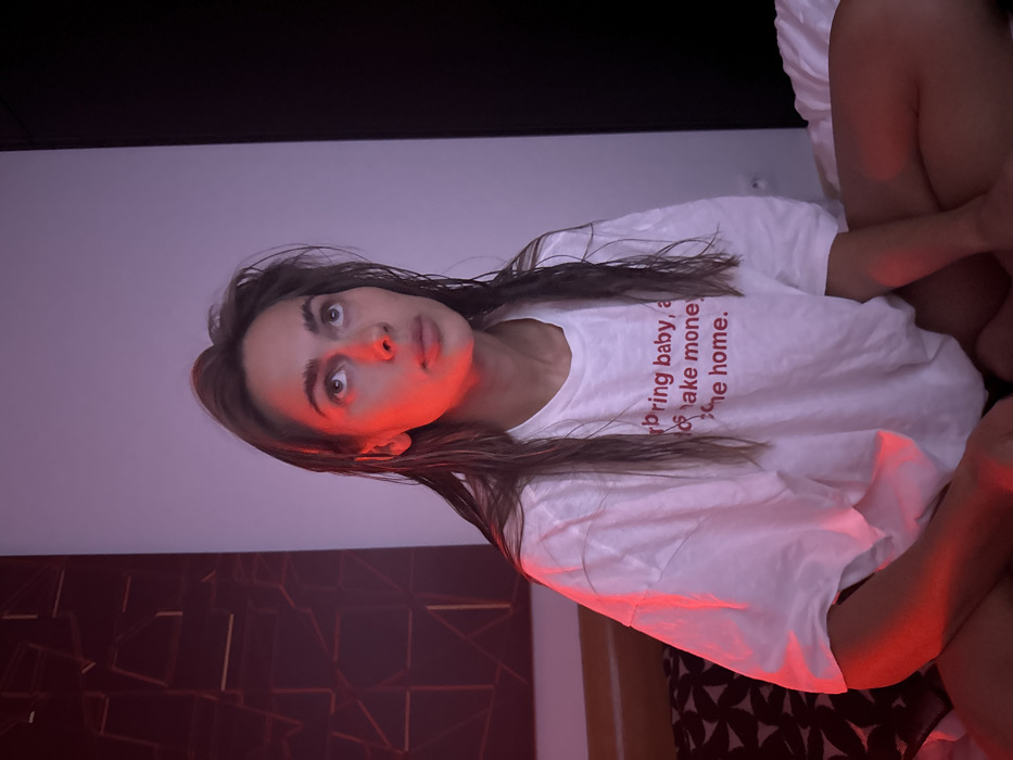 A woman with brown hair and wide eyes looks upward, illuminated by a reddish light on her face. She wears a white t-shirt with red text. Another person's legs are in the upper right, on white bedding. The background includes a gray wall, a dark shelf or headboard above, and a patterned brown and black wall design. The image is shot from a low angle and slightly tilted.