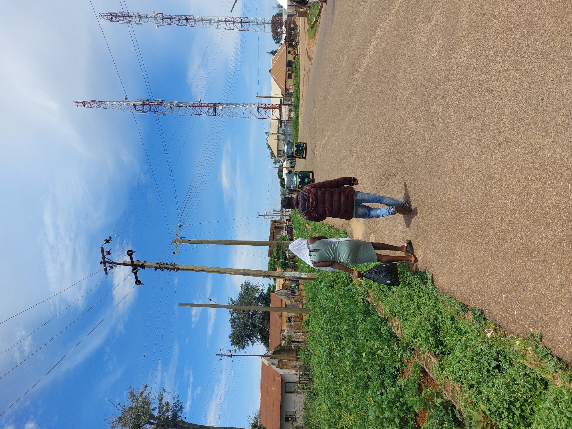The image, captured in Barakin, Nigeria, depicts a roadside scene under a clear blue sky with wispy white clouds. Two individuals are walking along a paved road, which appears to be the main thoroughfare. The person in front, a woman, is wearing a light green dress and a white headscarf, and is carrying a black bag. She is walking away from the camera, with her back slightly turned. Behind her, a man in a dark maroon jacket and blue jeans is also walking away from the camera.
The setting is a semi-rural or suburban area, with houses and other structures visible in the background. Several tall telecommunication towers dominate the skyline on the left side of the image, indicating a developed infrastructure. Power lines run across the frame, supported by wooden utility poles. Lush green vegetation lines the side of the road, suggesting a warm and possibly humid climate. Two three-wheeled vehicles, commonly known as "Keke Napep" or "Marwa," are parked or moving on the road in the distance.
The image appears to be taken during daylight, likely late morning or early afternoon, given the bright sunlight and shadows. There are no explicit indications of specific emotions, but the individuals seem to be walking at a casual pace, suggesting a normal daily activity. The scene evokes a sense of everyday life in a Nigerian town. No visible text is clearly discernible in the image.