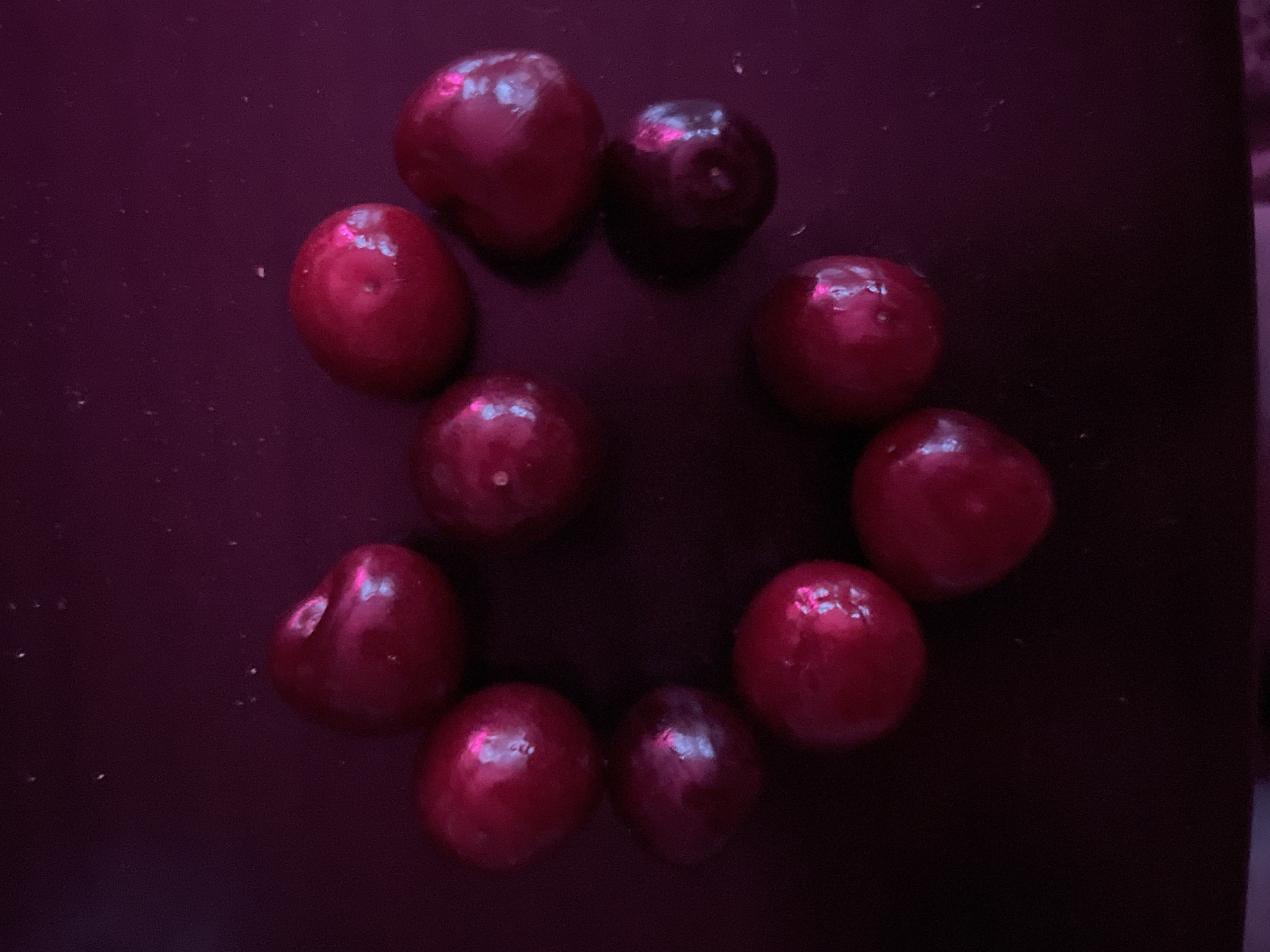 A close-up, overhead shot shows several shiny, dark red cherries arranged in a heart shape on a dark, glossy surface. The lighting is dim and casts a pinkish-purple hue over the scene, creating reflections on the cherries and the surface. There is no discernible background or environmental context that would place this image in Vyborg, Russia, or any specific city. The focus is entirely on the arrangement of the cherries.