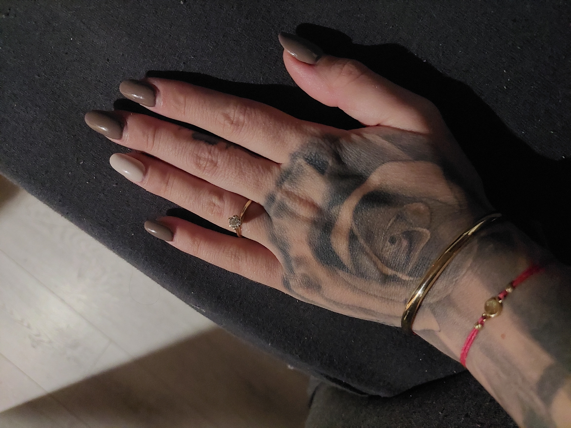 A close-up, slightly high-angle shot reveals a tattooed hand adorned with jewelry resting on a dark, textured surface. The hand is light-skinned with a large, faded black-ink tattoo depicting a rose on the back of the hand extending to the forearm. The fingernails are painted in varying shades of brown except the ring finger, which is painted a light, off-white color. A gold-colored ring with a central stone sits on the ring finger. The wrist is adorned with two bracelets: a gold-colored bangle and a thin red string bracelet with a gold-colored charm. The background suggests a dark, possibly fabric-covered surface. There are no indications of the city of Plovdiv, Bulgaria, or any identifiable landmarks or objects associated with the location. The scene contains no visible people beyond the depicted hand and arm.