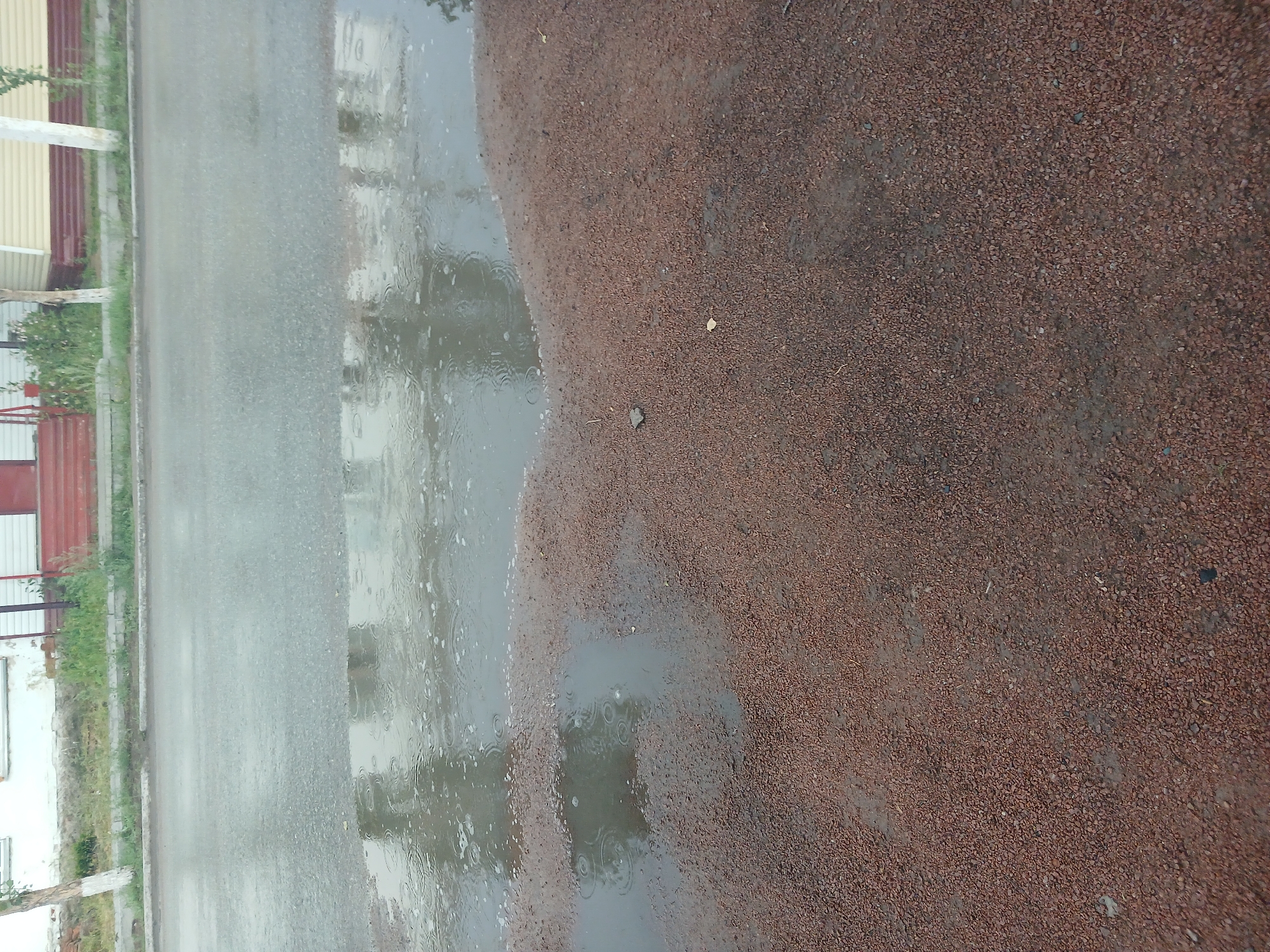 The image depicts a street scene in Makinsk, Kazakhstan, after rainfall. A large puddle of water occupies the center and left portions of the frame, reflecting the overcast sky and the buildings lining the street. The street itself is wet and appears to be asphalt. The foreground consists of a path covered in small, reddish-brown gravel, which is also damp. Buildings, possibly residential, are visible in the background with elements of their facades, including windows and doors, and some greenery. The overall impression is one of a quiet, damp day in the city.