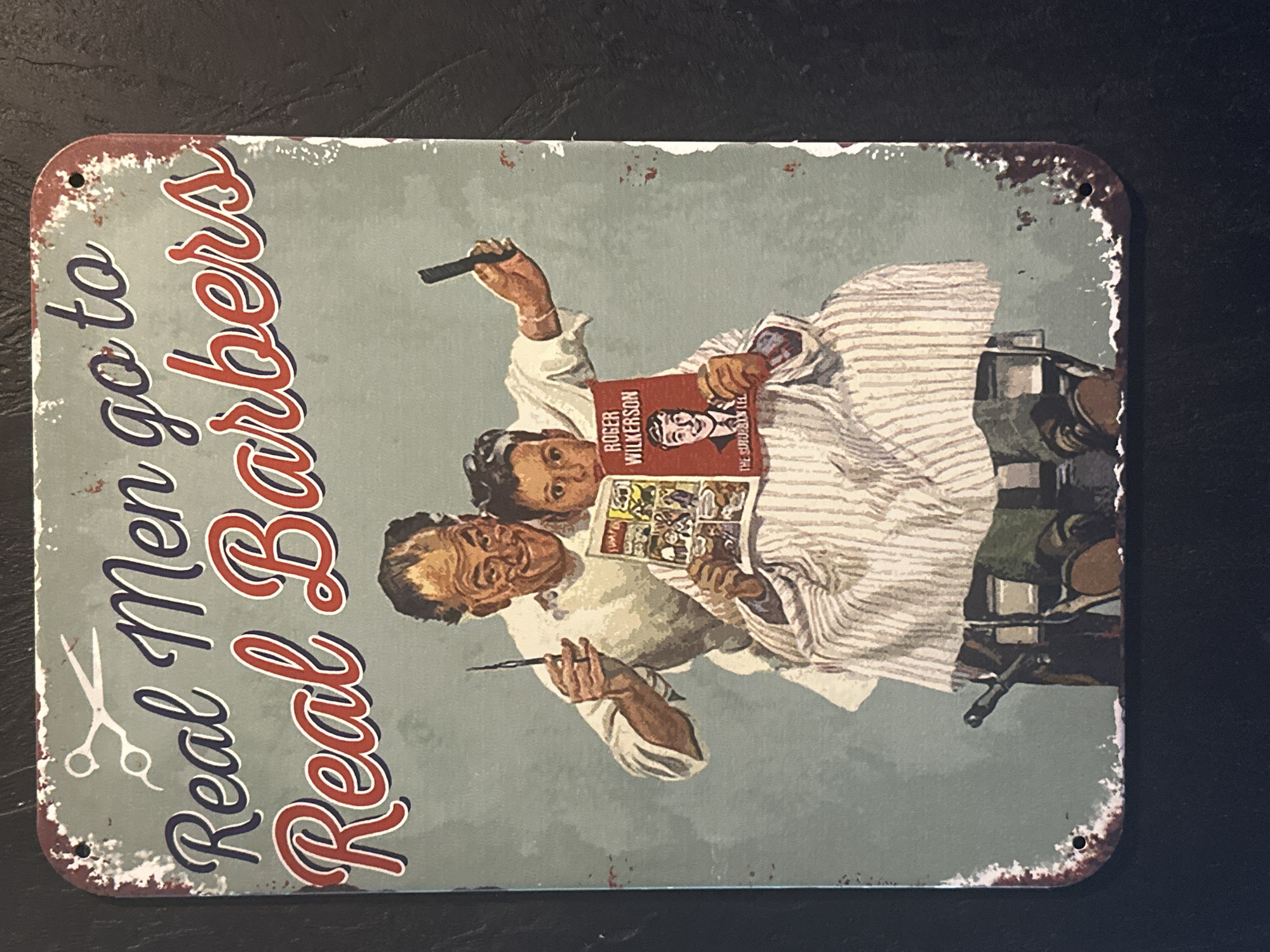 The image depicts a rectangular tin sign with a weathered, vintage appearance. The sign features a faded light blue-gray background. On the left side, in large red and navy cursive lettering outlined in white, it reads "Real Men go to Real Barbers". A pair of scissors is illustrated next to the text.
The right side of the sign shows a vintage illustration of a barber and his client. The barber is shown holding a straight razor. The client sits in a barber chair covered in a striped barber cape and is reading a comic book. He is also holding a book titled "Roger Wilkerson". The illustration has a slightly distressed, aged aesthetic.
The sign has a rusted-look trim with small holes in each corner, presumably for mounting. The environment appears to be a dark, neutral surface upon which the sign rests.
There is no information to suggest that this image is located in the city of Selyatino, Russia.