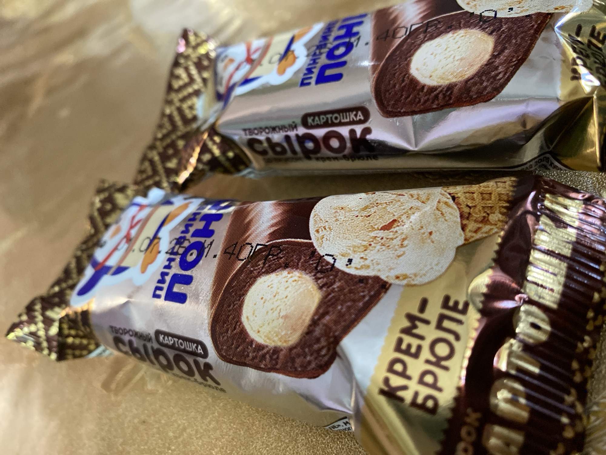 The image shows two individually wrapped ice cream bars with foil packaging, placed on a textured golden surface. The packaging is predominantly silver with accents of gold and blue. One ice cream bar on the left has visible text in Russian, including "КРЕМ-БРЮЛЕ" (Crème brûlée) in large, bold letters. The other ice cream bar on the right also has Russian text. Both packages feature cartoonish illustrations of animals wearing hats. The ice cream bars themselves appear to be chocolate-covered with a creamy white center, and one of the bars is partially unwrapped to reveal a scoop of vanilla ice cream on a waffle cone. The lighting suggests an indoor setting. There is no identifiable context for the city of Maykop, Russia.