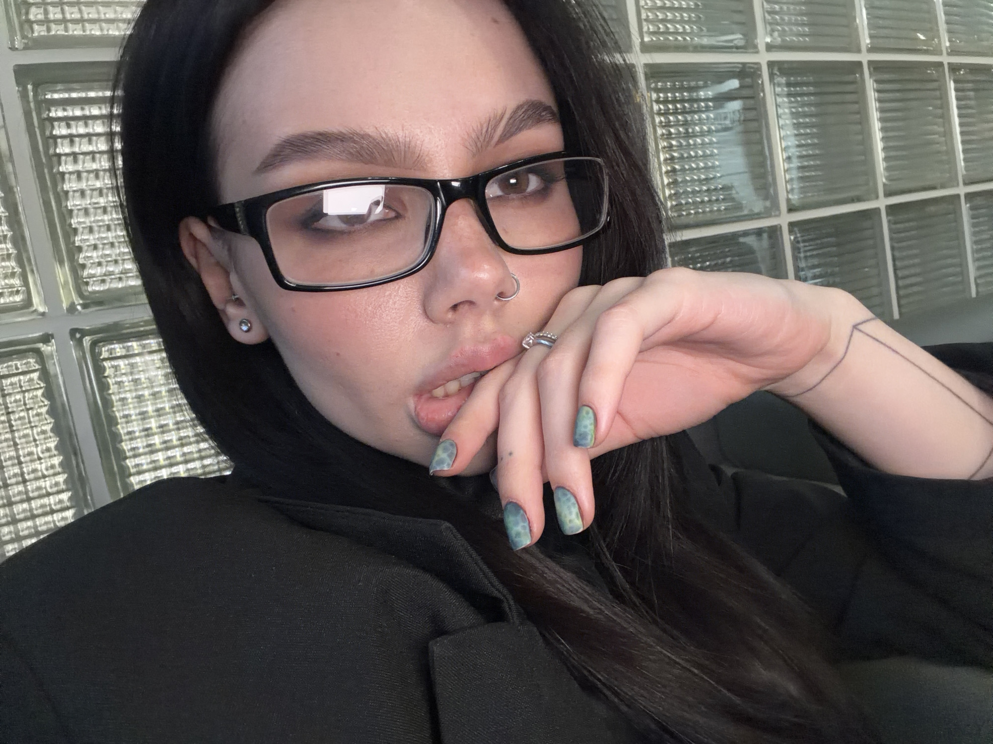 A woman with dark hair and glasses poses for a selfie indoors against a backdrop of glass blocks. She wears a black blazer and has a nose ring. Her nails are painted with a blue and green pattern. She holds her hand near her mouth, partially covering it. A ring adorns her finger, and a subtle tattoo is visible on her arm. The location cannot be ascertained to be in Moscow, Russia.