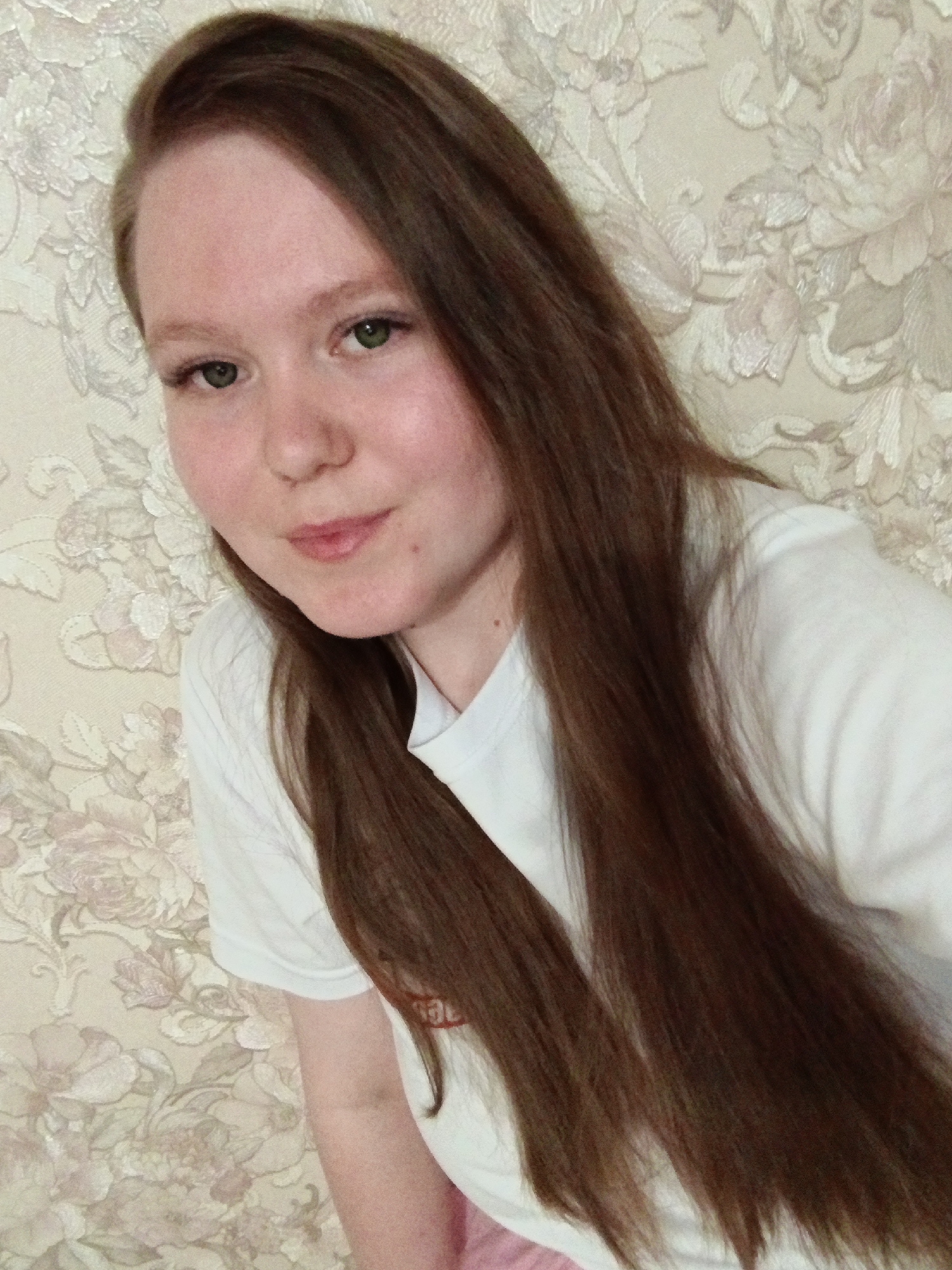 A young woman with long brown hair and green eyes is posing for a selfie. She is wearing a white t-shirt. The background is a wall covered in a floral wallpaper pattern. The location is likely indoors in Chaikovsky, Russia.