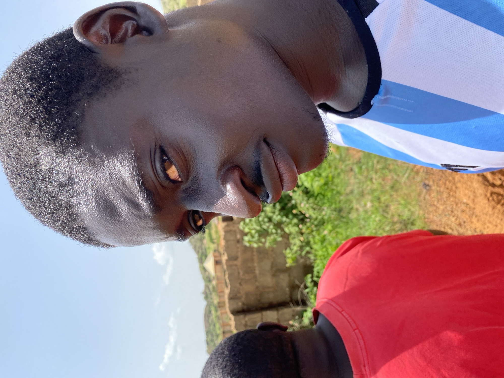 The image is a close-up, outdoor portrait of two young Black males in Barakin Angundi, Nigeria. The primary subject, in the foreground and facing the camera, is a male with dark skin and short, dark, curly hair. He is wearing a blue and white striped jersey with a black neckline. His head is tilted slightly, and he is looking towards the camera with a subtle smile. His right eye is visible, and the light reflects off his forehead and cheek. Behind him and to the left, a second male is seen from the back, wearing a bright red t-shirt. The background consists of a low, reddish-brown brick wall, lush green foliage, and a hazy sky with some clouds. The ground appears to be a mix of reddish-brown soil and dry grass. The overall impression is a casual, sunny outdoor setting.