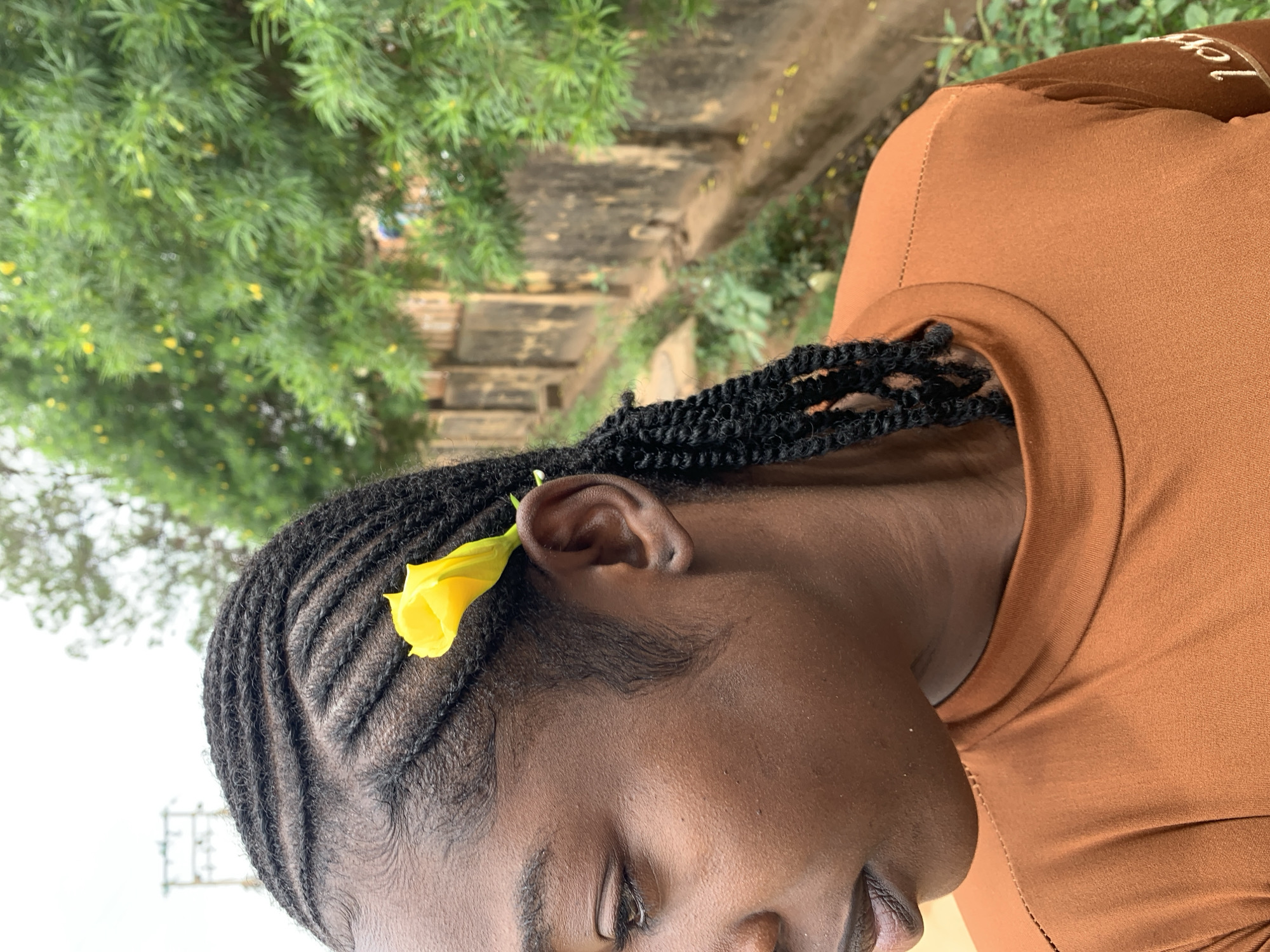 The image displays a close-up, left-profile portrait of a female individual located in the city of Rafin Par, Nigeria. Her hair is styled in distinct black cornrows on the scalp, transitioning into twisted braids that extend down the side. A single, vibrant yellow flower is positioned behind her left ear. The individual is wearing a long-sleeved, rust-orange colored shirt with a crew neck, featuring a horizontal stitching pattern on the left shoulder. Her head is angled slightly to the left, and her gaze is directed downwards. The background is out of focus, revealing an overcast sky and utility poles in the upper left quadrant. The right side of the background consists of dense green foliage containing small yellow flowers, situated above a weathered, dark-colored concrete wall and a narrow, unpaved drainage channel.