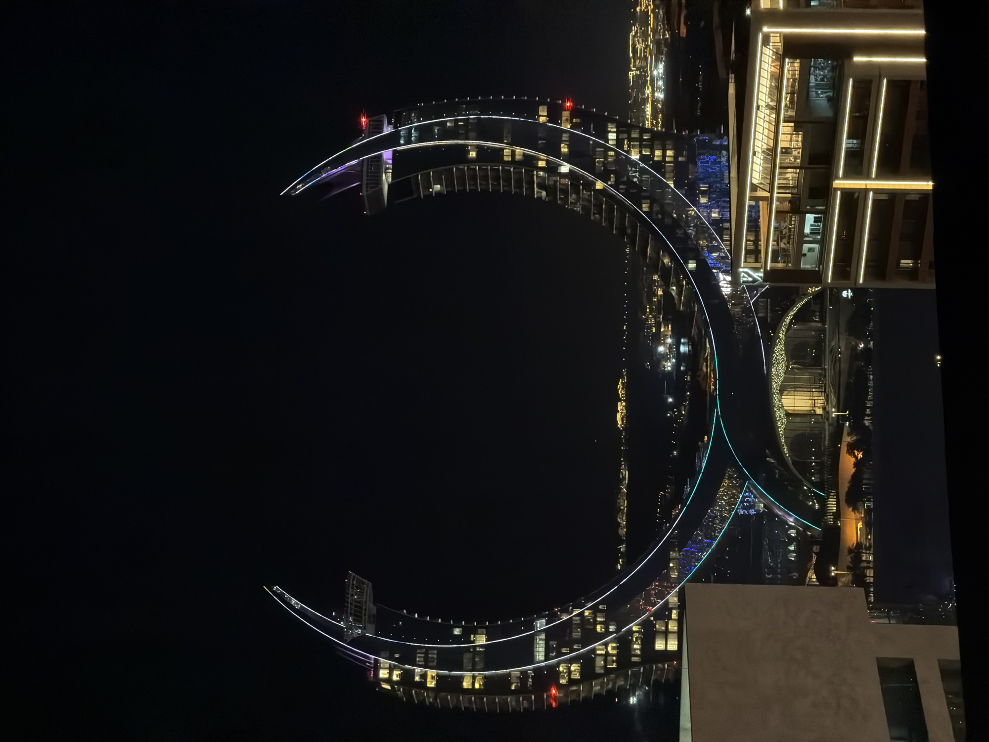 The image shows a nighttime aerial view of a city at night, with a focus on a large, illuminated architectural structure resembling a crescent moon or an incomplete circle. The structure is outlined with bright white lights, and there are also colorful lights, including blue and green, accentuating its curves and parts of the surrounding cityscape. The city is filled with numerous lights from buildings, indicating it is a densely populated urban area. Reflections of the lights can be seen on a body of water below, suggesting the structure might be near or over water.
The perspective is from a high vantage point, looking down and across the scene. The architecture of the illuminated structure is modern and sleek. Some buildings on the right side of the image are also well-lit, with horizontal bands of light. The overall scene is one of urban development and modern design at night. The mood is illuminated and somewhat futuristic due to the prominent lighting. There is no visible text in the image. The weather appears to be clear, as the lights are sharp and the reflections are well-defined. The time of day is clearly night.