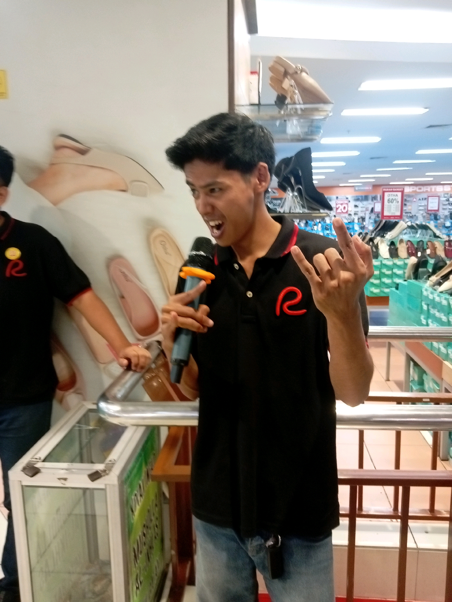 In Batam, Indonesia, a young man stands in a shoe store, holding a microphone and making a rock-and-roll hand gesture. He is wearing a black polo shirt with a red "R" logo and blue jeans. The background shows shelves of shoes and a price tag indicating a 60% discount. The man's expression is excited, suggesting he is either singing or addressing a crowd. The store's bright lighting and displays suggest it is a busy commercial environment.