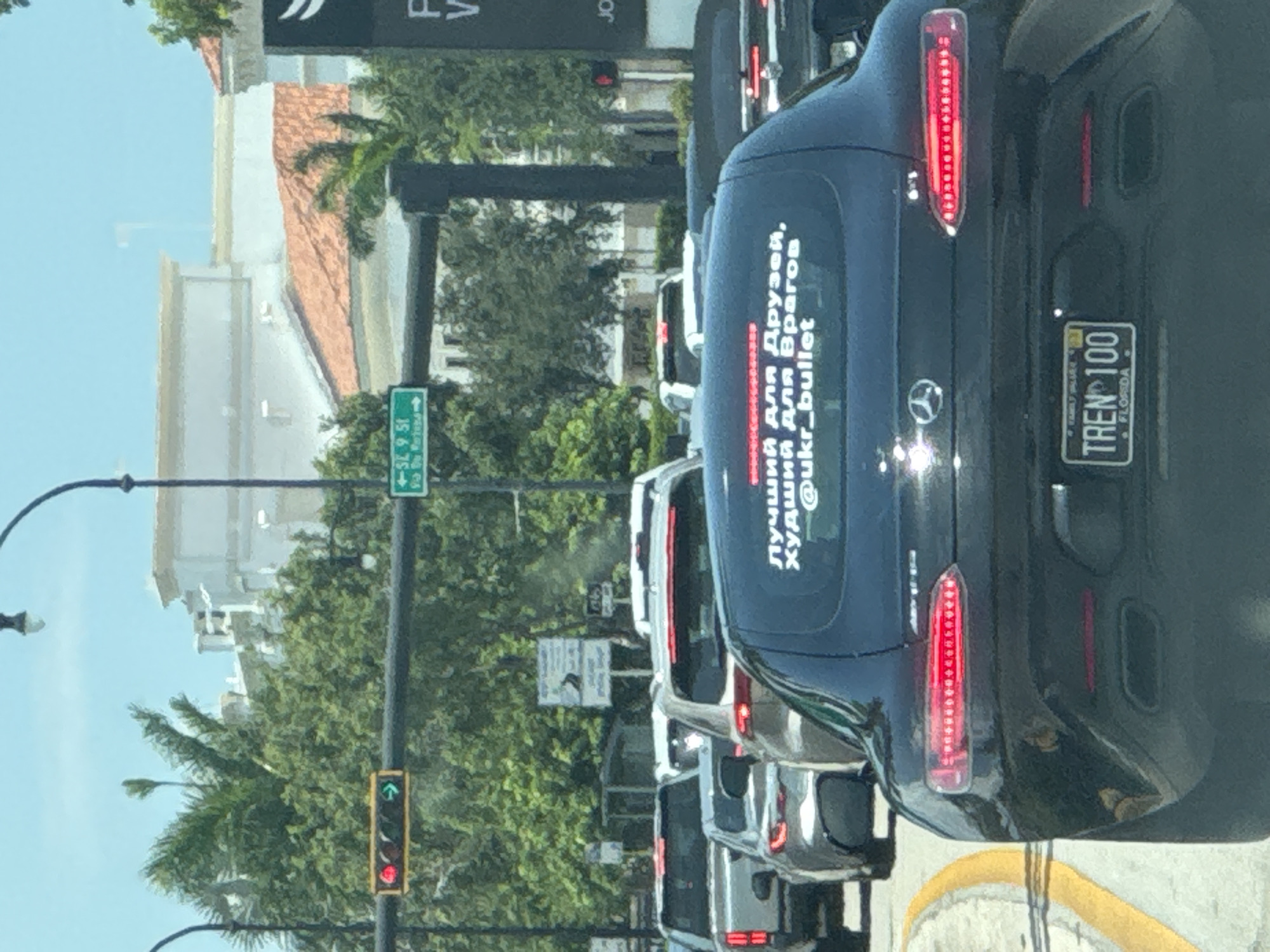 In Hallandale Beach, United States of America, a black Mercedes-Benz AMG GT coupe is stopped in traffic, facing away from the viewer. The car has a "TREN 100" license plate that reads "FAMILY VALUE" and "FLORIDA". A white sticker on the rear window contains Cyrillic text that translates to "Best for Friends, Worst for Enemies" followed by "@ukr_bullet". Several other vehicles are also stopped in traffic, including a silver SUV and a black car. Traffic lights are visible, with one showing red and green arrows. Green street signs with white text are also present. The environment is an urban street with trees and buildings in the background.