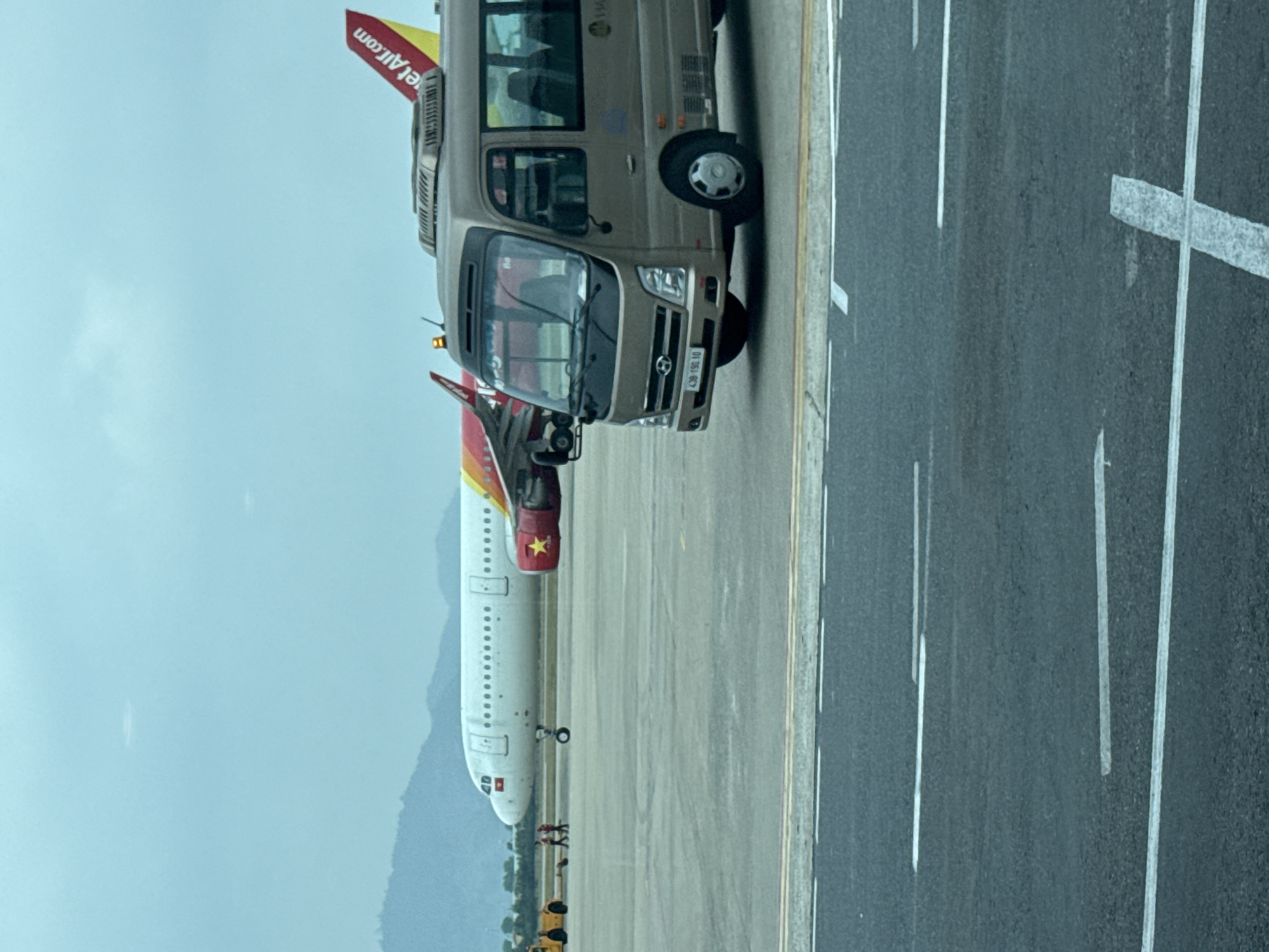 The image shows a VietJet Air plane and a shuttle bus on an airport tarmac. The plane has a red and yellow tail with "VietJetAir.com" written on it. The shuttle bus is beige. Markings on the tarmac include white lines. A Vietnamese flag is visible on the plane. Several people are visible near the front of the airplane. The airport is located in Đà Nẵng, Vietnam.