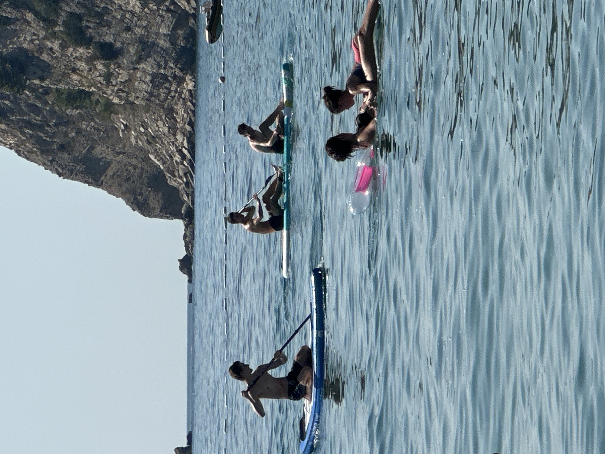 The image shows a sunny day at the beach in Đurovići, Montenegro. In the foreground, two young girls are playing in the water on inflatable toys. Behind them, three young men are paddleboarding. The man on the left is kneeling and paddling, while the other two are sitting on their boards. In the background, a large rocky cliff rises from the sea. The water is a clear blue, and the sky is a pale blue. The scene is peaceful and idyllic, capturing a typical summer day at the beach.