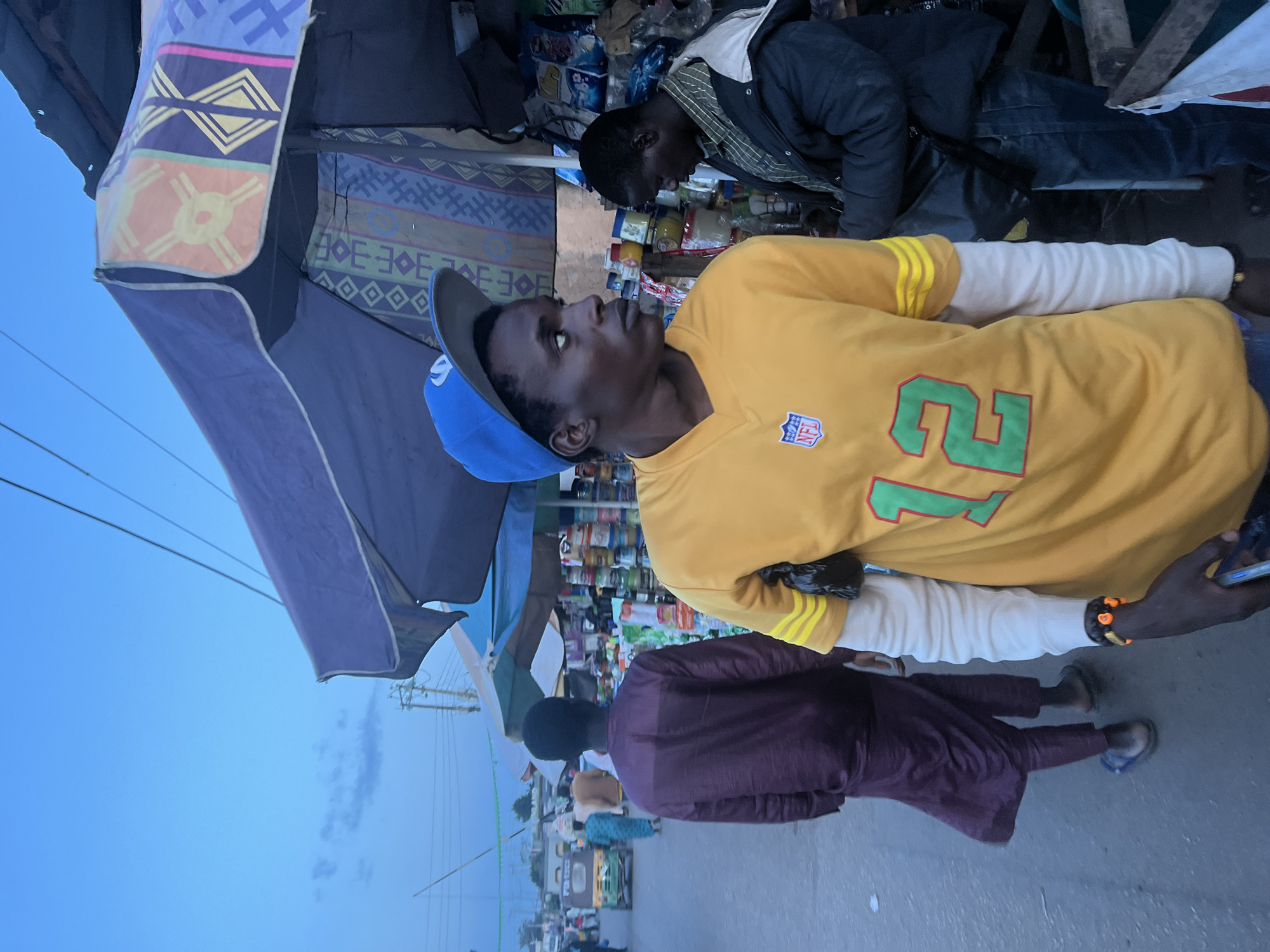 A young Black man wearing a blue baseball cap and a yellow long-sleeved shirt with the number "12" on it walks past a market stall. He is looking to his right, away from the camera. Behind him, another Black man in a dark jacket and jeans appears to be browsing the stall. The market stall is under a large purple umbrella with colorful geometric patterns. The background shows a street scene with other people, vehicles, and stalls, suggesting a busy urban environment. The sky is a pale blue, indicating it might be early morning or late afternoon.