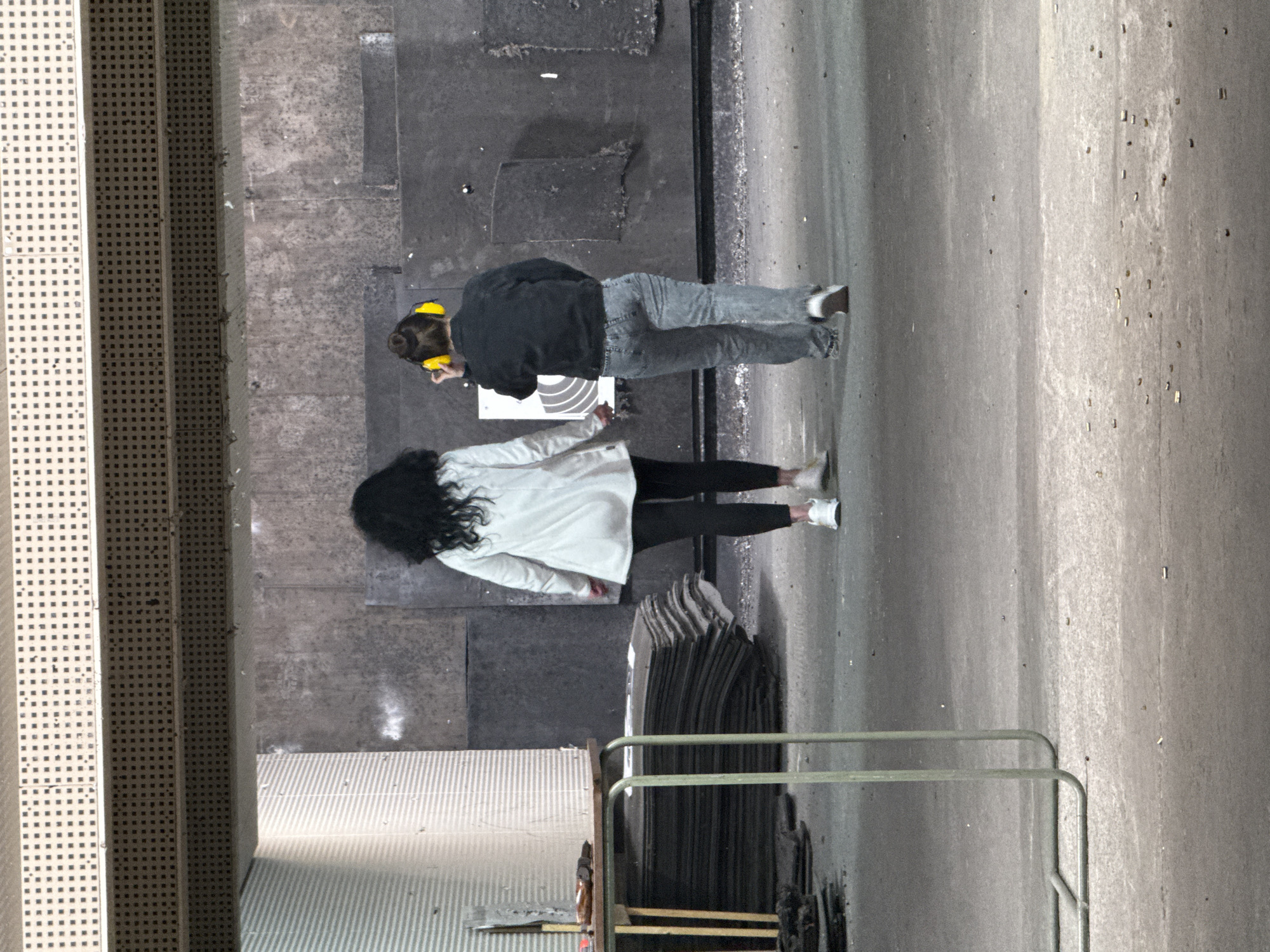 This is an overhead shot of two individuals, likely women, standing in what appears to be an indoor or covered industrial space, possibly a shooting range, given the ear protection worn by one person. The image is taken from a slightly elevated position, looking down.
The person further from the camera has dark hair tied in a bun, adorned with bright yellow ear protection or headphones. They are wearing a dark jacket and light gray jeans. They are looking down towards a white, circular target-like object placed on the ground in front of them.
The second person, closer to the camera, has long, dark, curly hair. They are dressed in a light-colored, possibly white, long coat or jacket and black pants, with white sneakers. They are standing near a stack of dark, flat, wave-edged sheets or panels.
The flooring is a concrete or asphalt-like surface with varying textures and shades of gray. To the left, there is a light-colored, perforated metal structure, possibly part of a staircase or platform. In the foreground, a light green metal railing is partially visible. The overall color palette is muted, dominated by grays, blacks, and whites, with the yellow ear protection providing a small color accent. There is no clearly legible text visible in the image. The setting is in Sofia, Bulgaria.