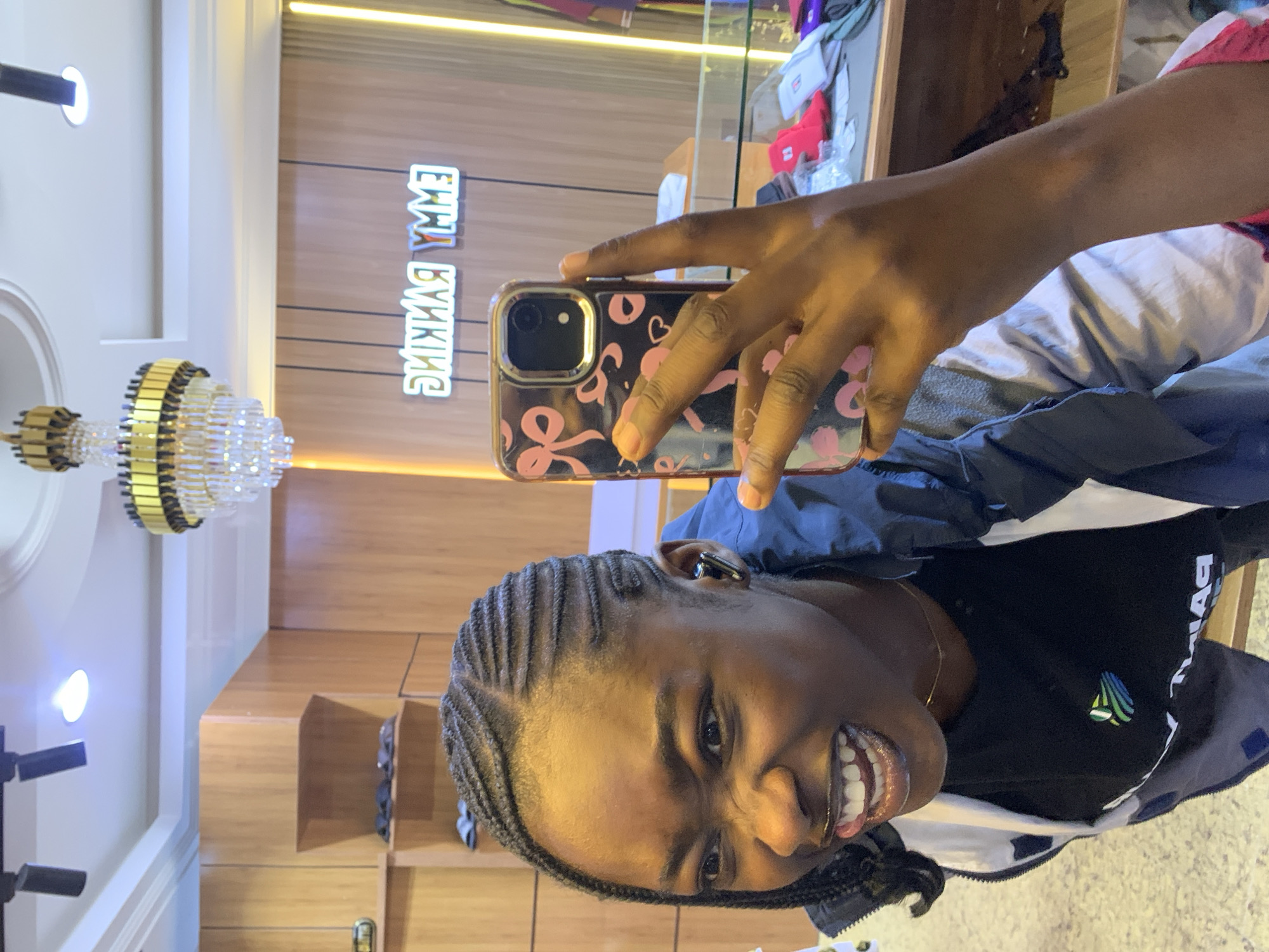 A young Black woman with braided hair takes a selfie in a store. She is wearing a black t-shirt and a blue and white jacket. She has a playful expression on her face, with her mouth open and tongue sticking out slightly. The background appears to be a retail environment, with wooden shelves and a neon sign that reads "EMMY RANKING". The store is likely located in Gura Top, Nigeria.
